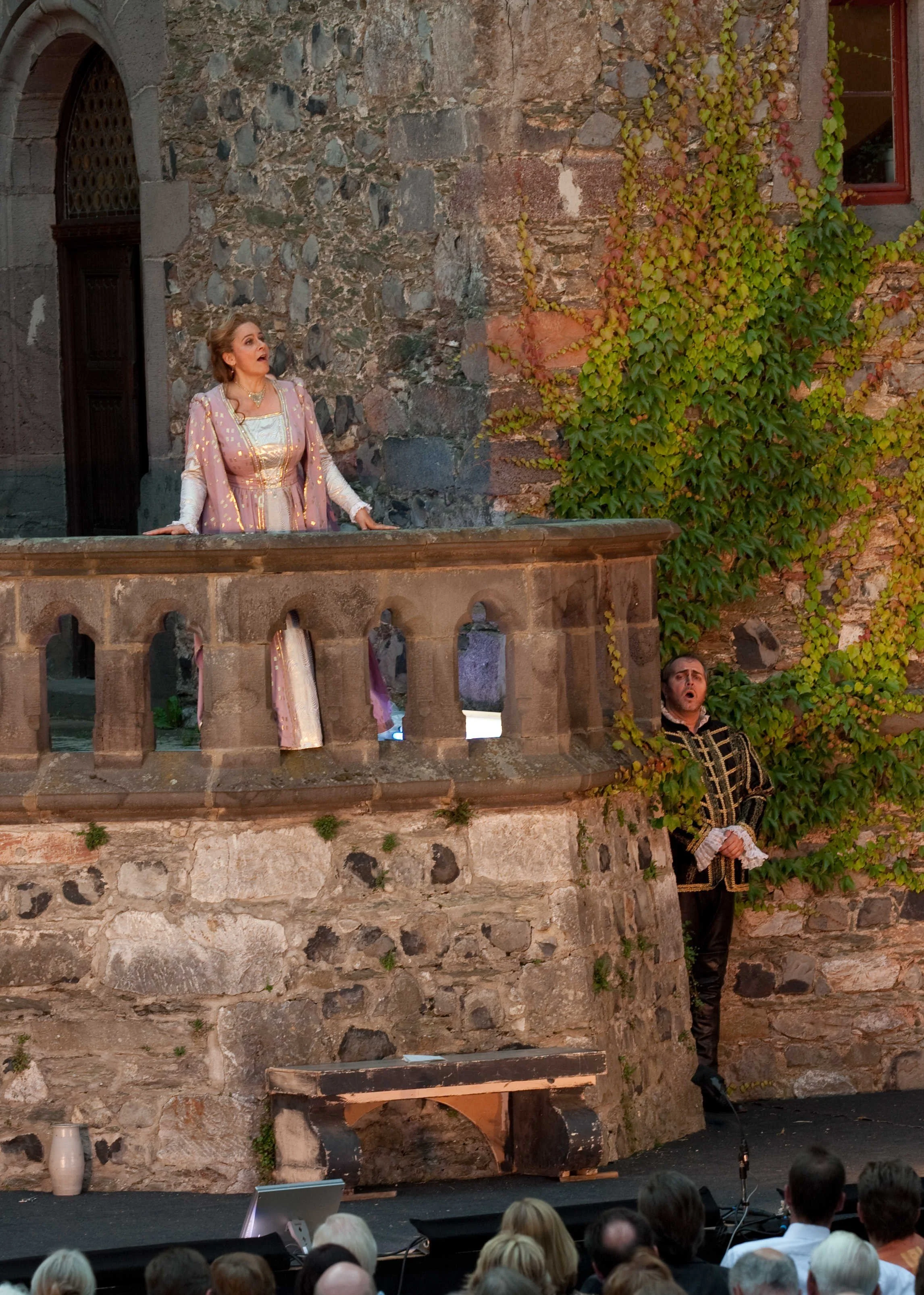 Scene from a theatrical performance on an outdoor stage resembling an old stone building with ivy, featuring a woman in period costume singing or acting, with an audience watching.