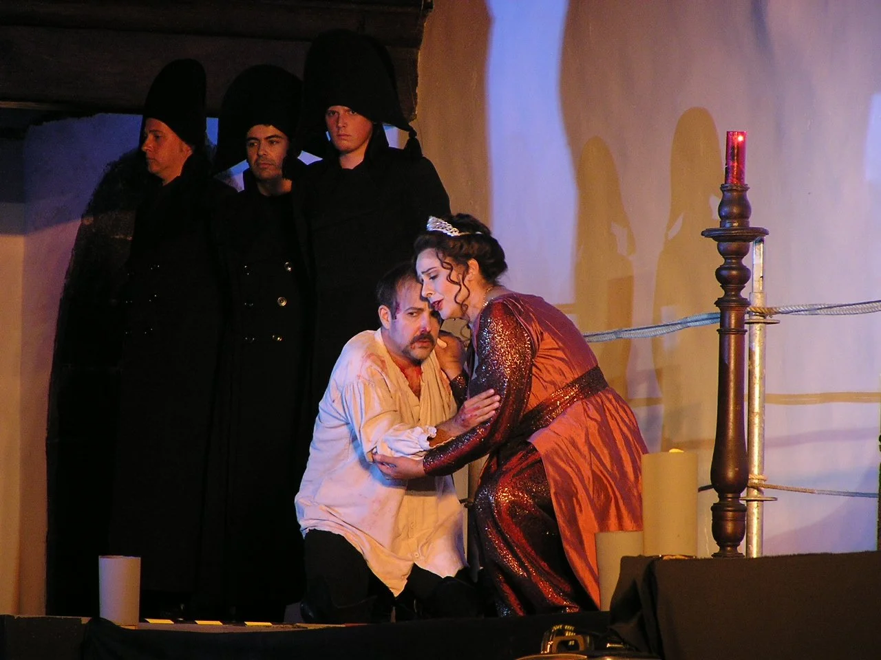 A theatrical scene with a man kneeling, looking distressed, while a woman in a shiny dress leans in, holding his arm. Three figures in dark robes and tall hats stand in the background, observing the scene.