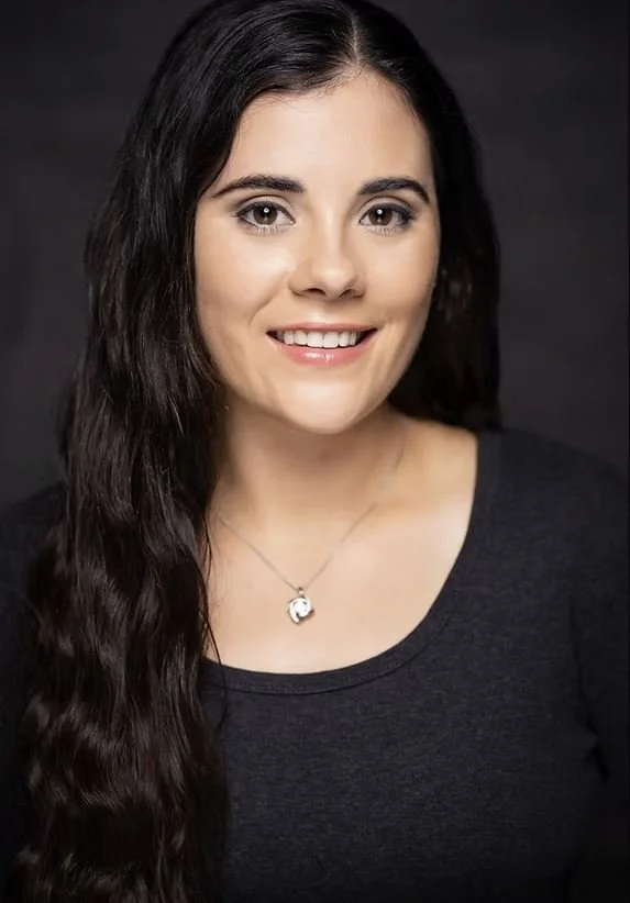 A woman with long, dark wavy hair smiling at the camera, wearing a dark top and a silver necklace with a pendant