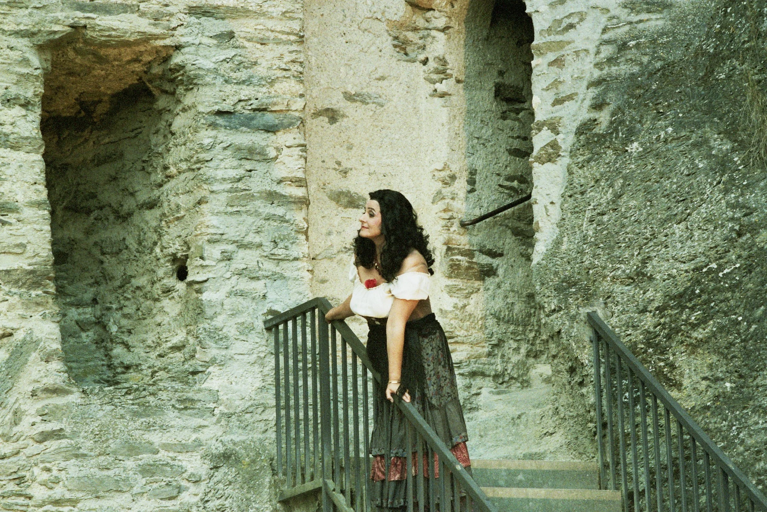 A woman with dark curly hair leaning on a railing at an ancient stone ruin or fortress, with textured stone walls behind her.