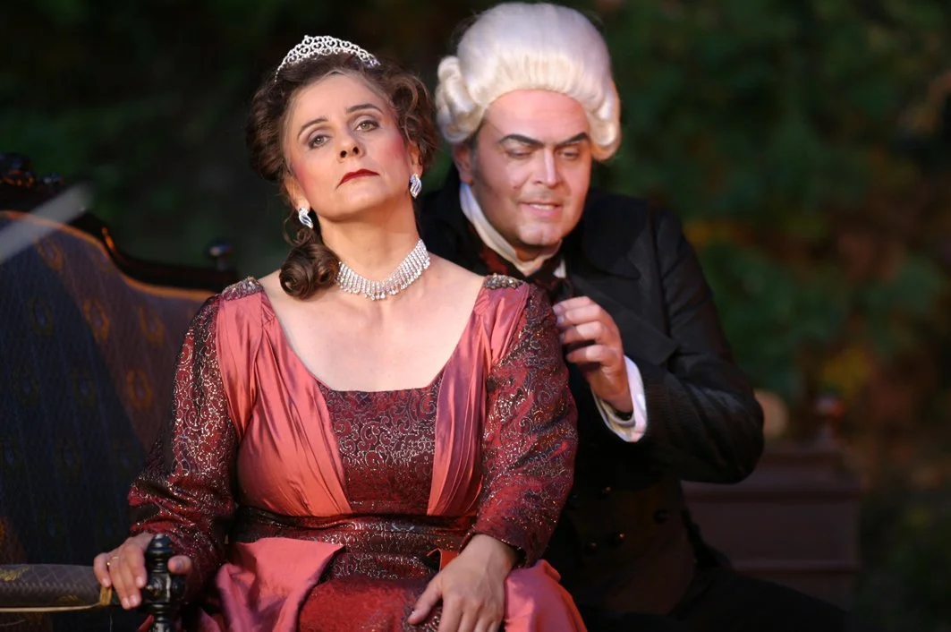 A woman dressed in period costume with jewelry and a tiara sitting on a chair, with a man in colonial attire standing beside her, in a theatrical setting.