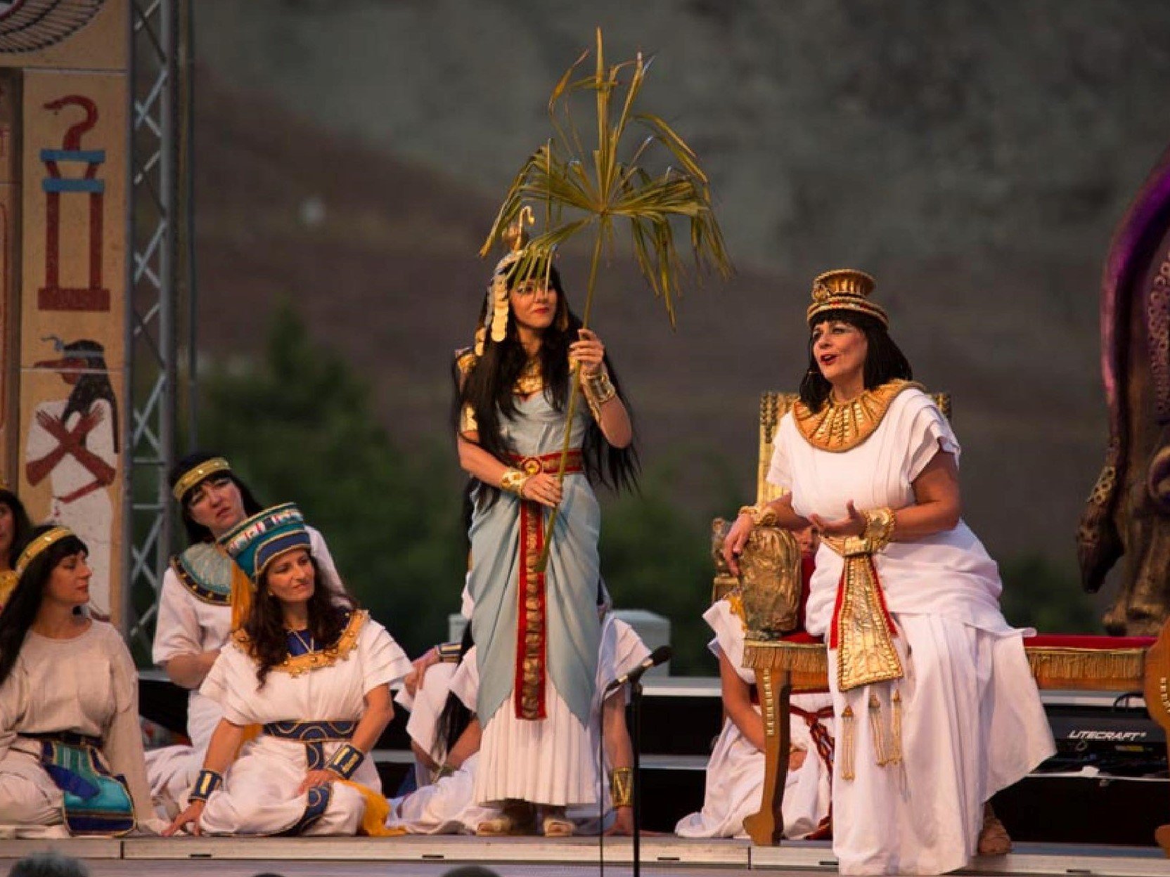 A group of people dressed in traditional Egyptian attire performing on stage with two women in the center, one standing holding a palm branch and the other seated with a gold or bronze frame around her neck, in front of a backdrop resembling ancient 
