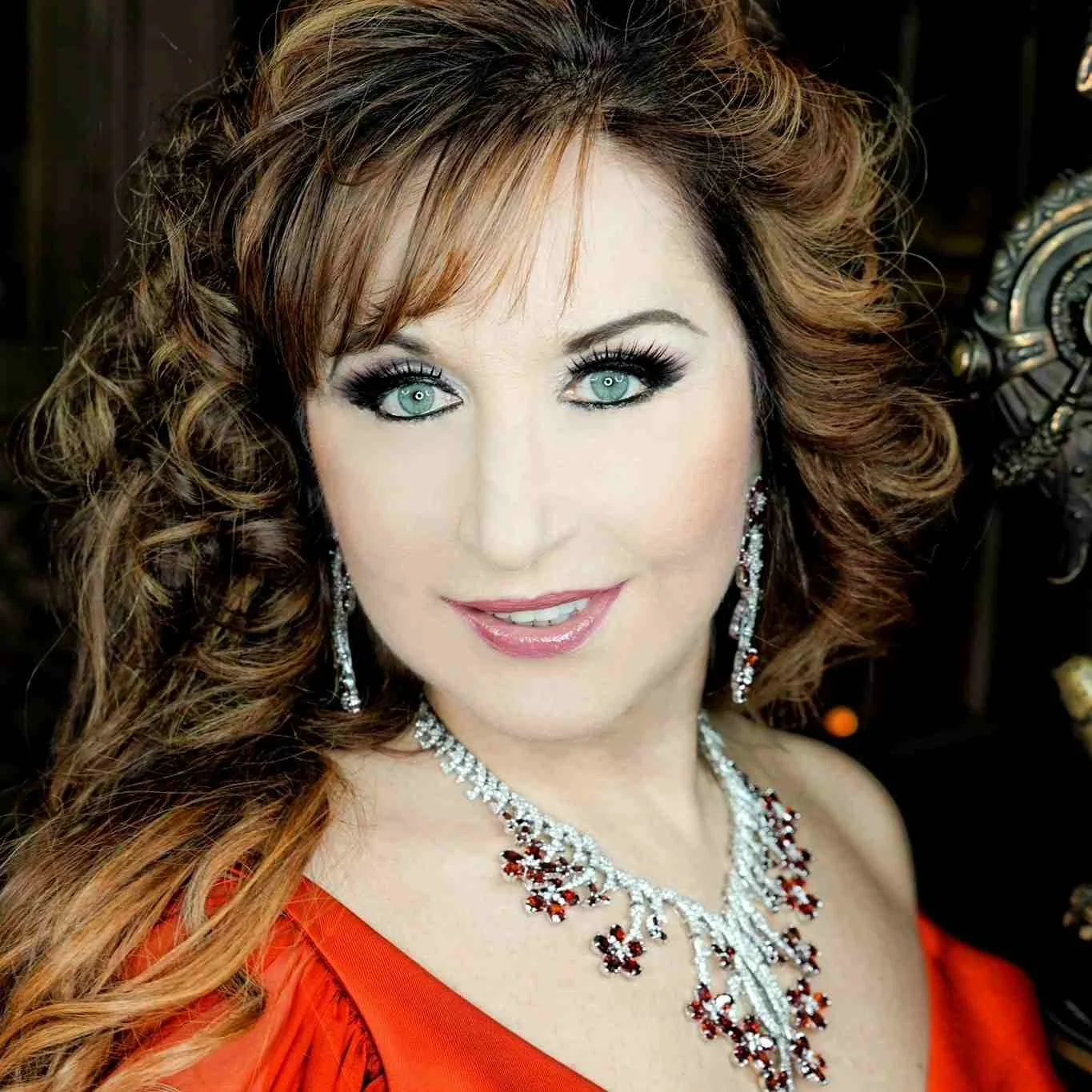 A woman with curly auburn hair, bright blue eyes, wearing a red dress, and adorned with silver and red jewelry, smiling at the camera.