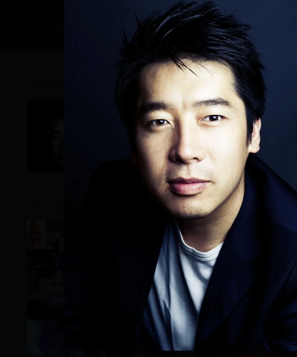 A portrait of an Asian man with dark hair, wearing a black blazer over a gray t-shirt, looking at the camera with a neutral expression against a dark background.
