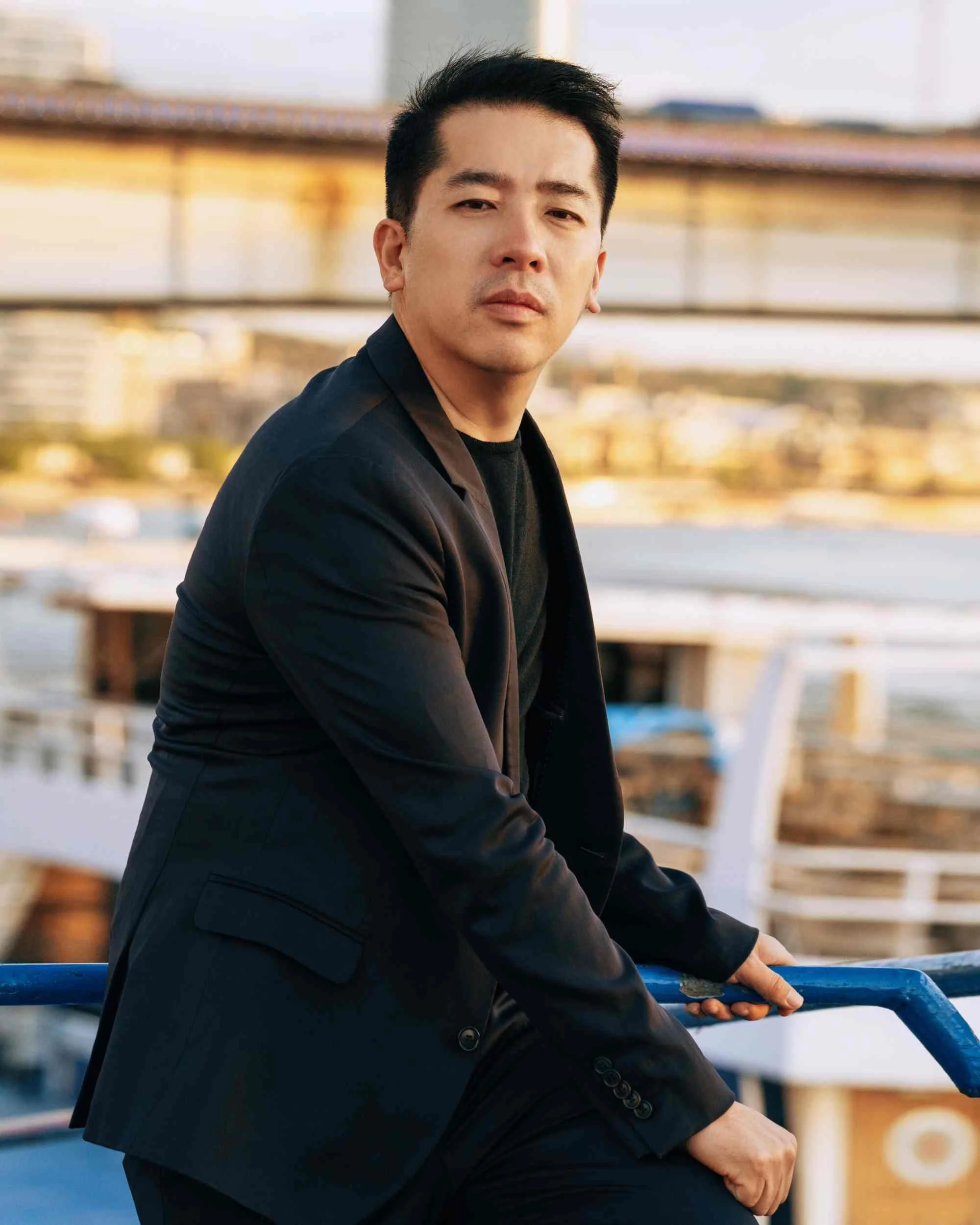 A man in a black suit sitting on a bench, holding onto a blue railing, in an outdoor setting with blurred buildings and structures in the background.