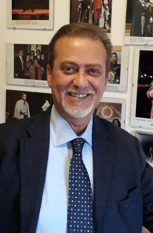 Smiling man with a beard and mustache in a suit, standing in front of a wall with framed photographs.