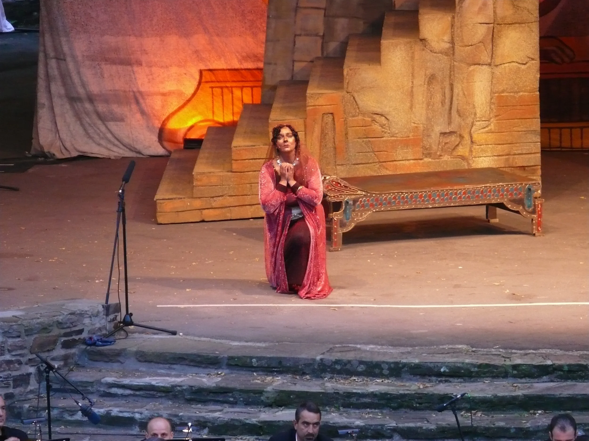 Woman in a red and pink dress standing on a stage with her hands clasped, behind her a large set of stone stairs and a mural painted on a backdrop, with microphone stands and part of an audience visible at the bottom.