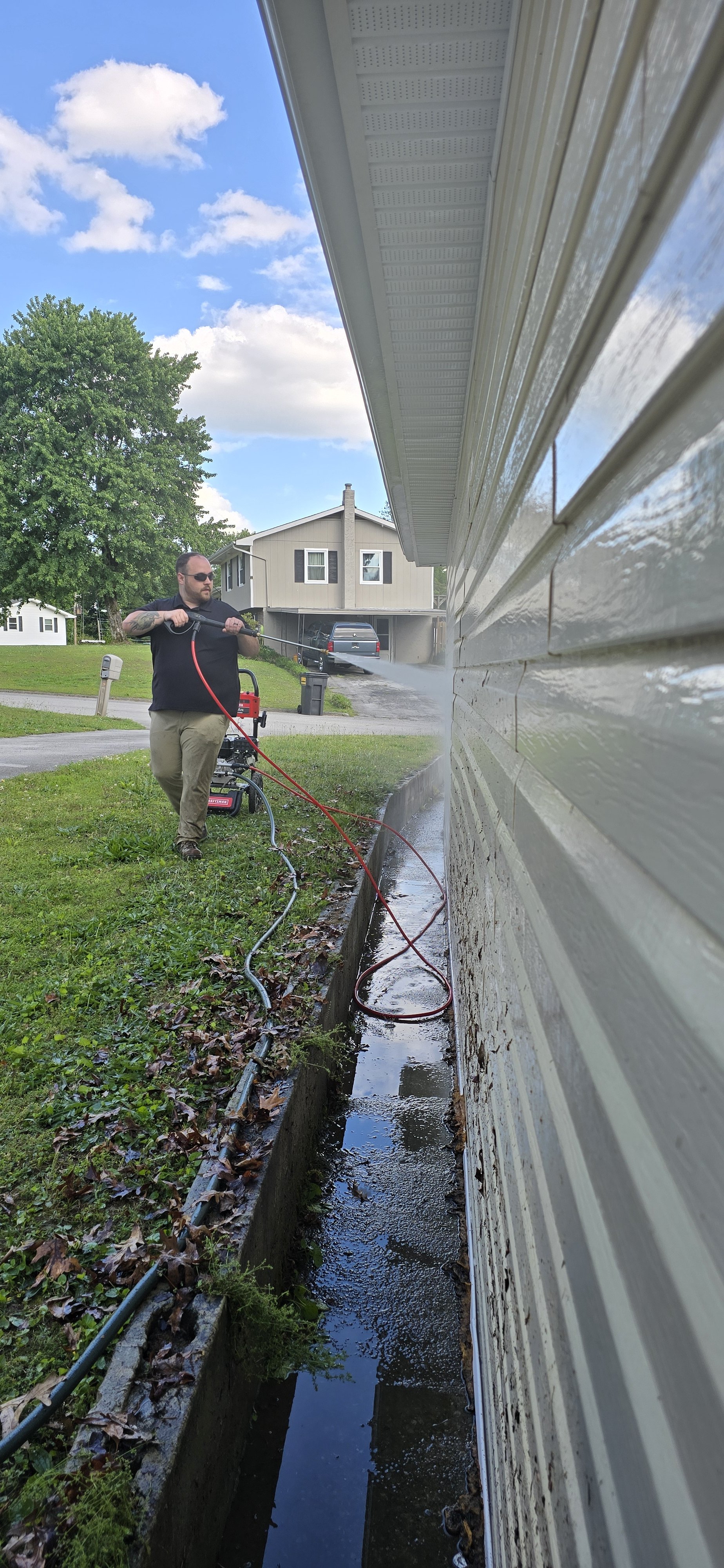 A man in sunglasses and dark clothing cleaning the side of a house with a pressure washer. Soft Washing in Hixson TN. Water is spraying from the nozzle onto the house's vinyl siding.