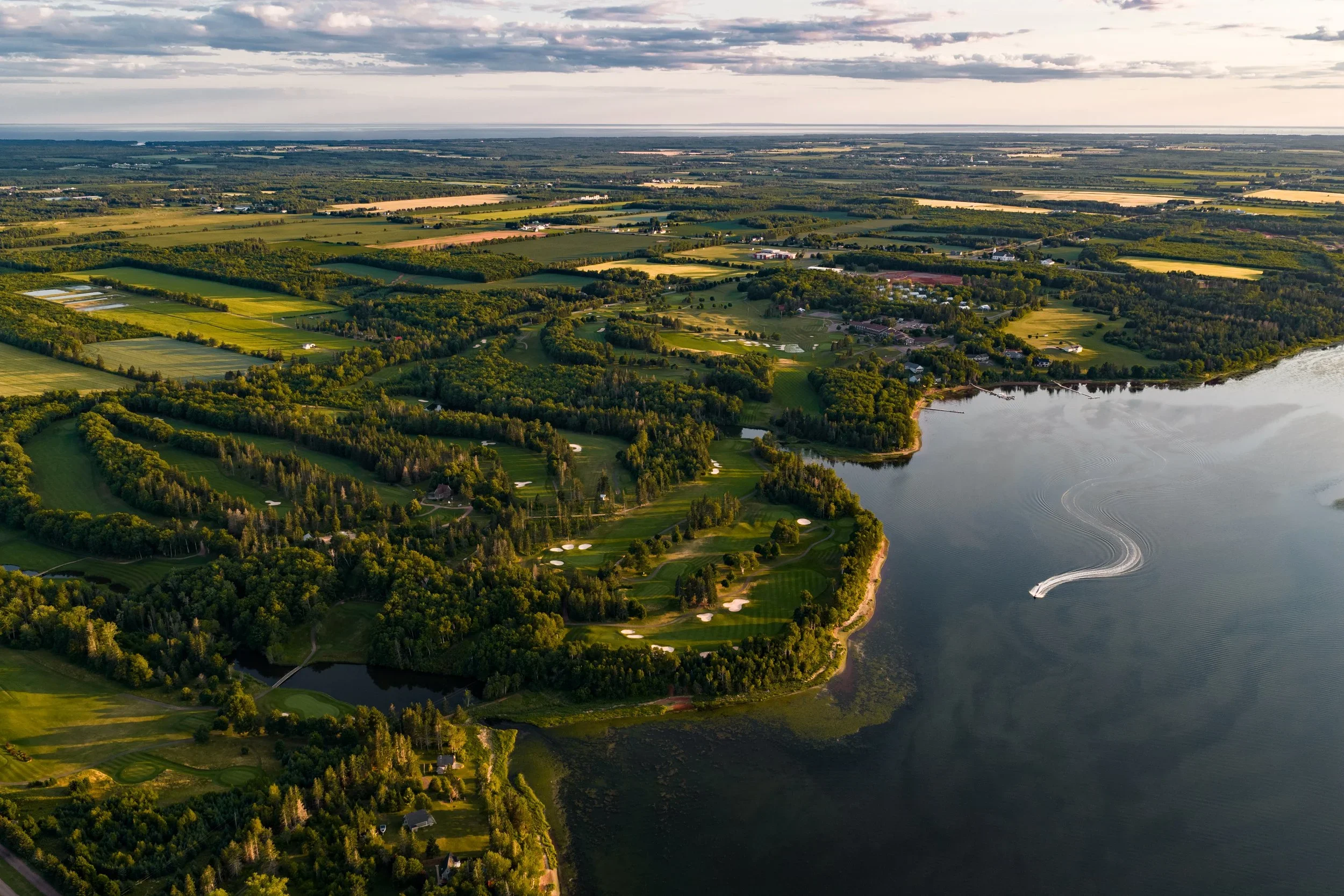 Aerial view of a golf course beside a large body of water, with lush green fairways, trees, and boats creating ripples on the water in the background.