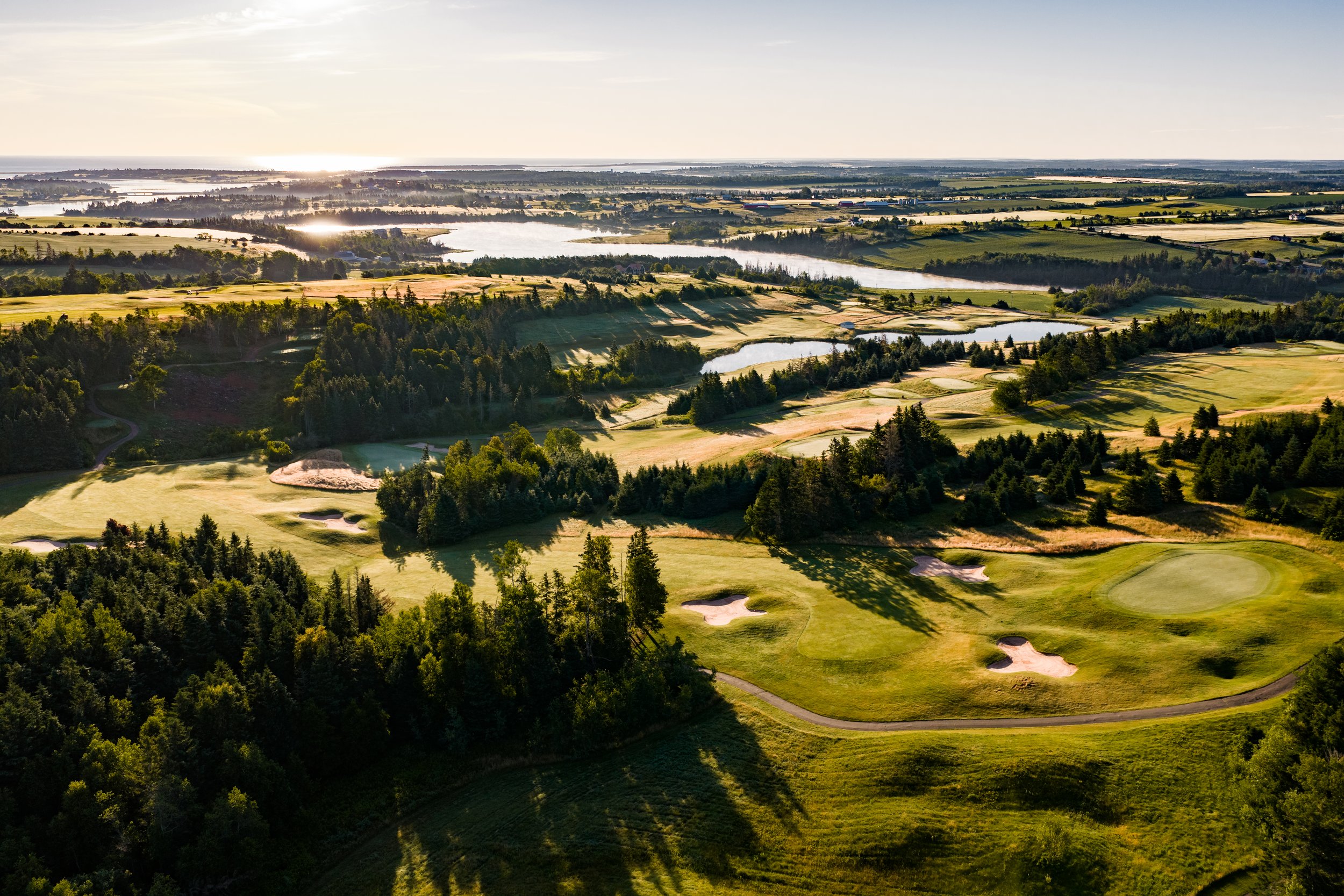 Aerial view of a golf course with lush green fairways, sand traps, surrounded by dense forest and water bodies, under a bright sky with sunlight reflecting off the water.