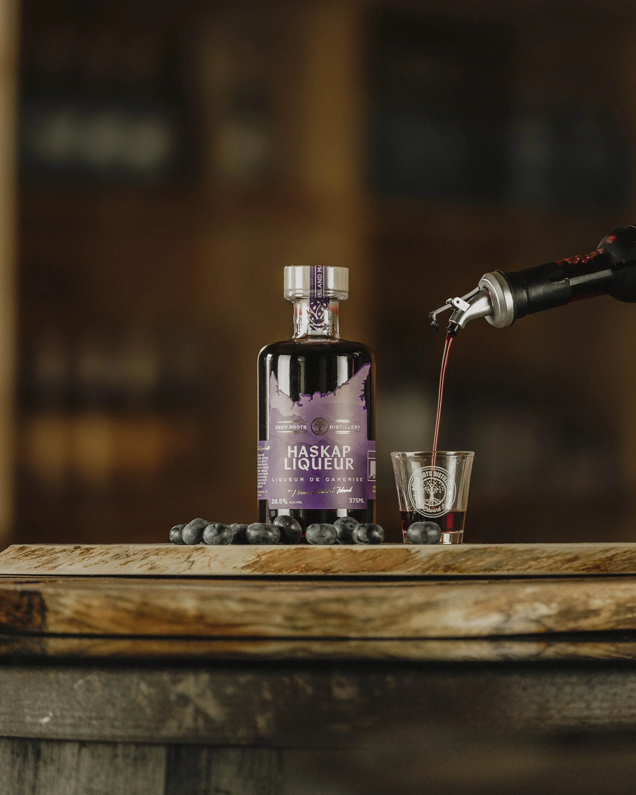 A bottle of Haskap Liqueur, blueberries, and a small glass filled with the liqueur on a wooden surface with blurred background.