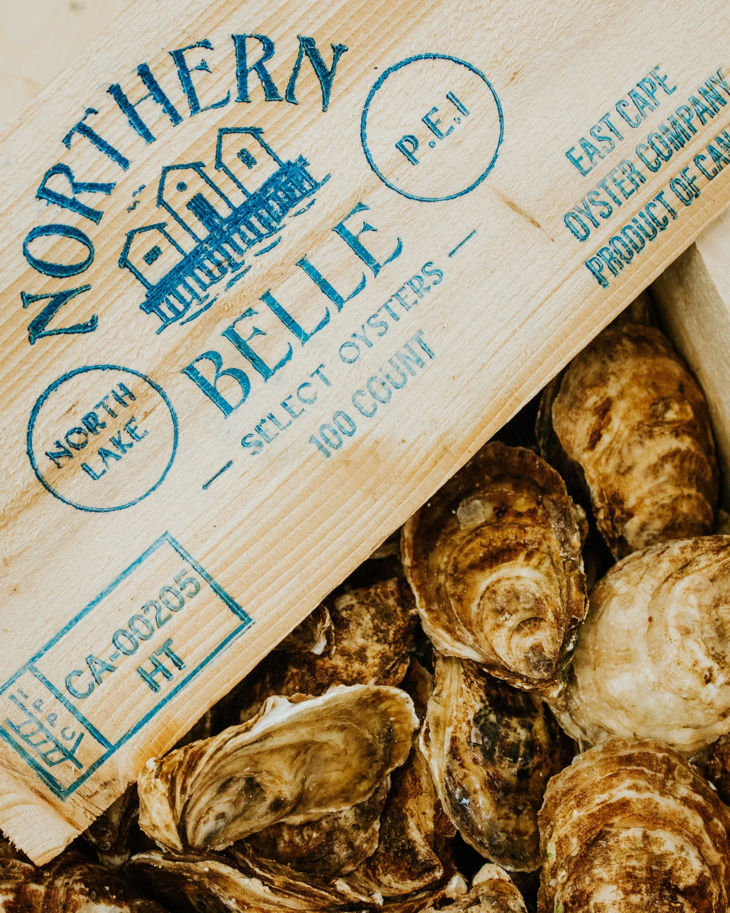 Close-up of a wooden crate label on a pile of oysters. The label reads 'ZOTI HENRY,' with a drawing of buildings, and mentions 'BELLE EAU' and 'EAST CAPE P.D.E' with text about the oyster company and location.