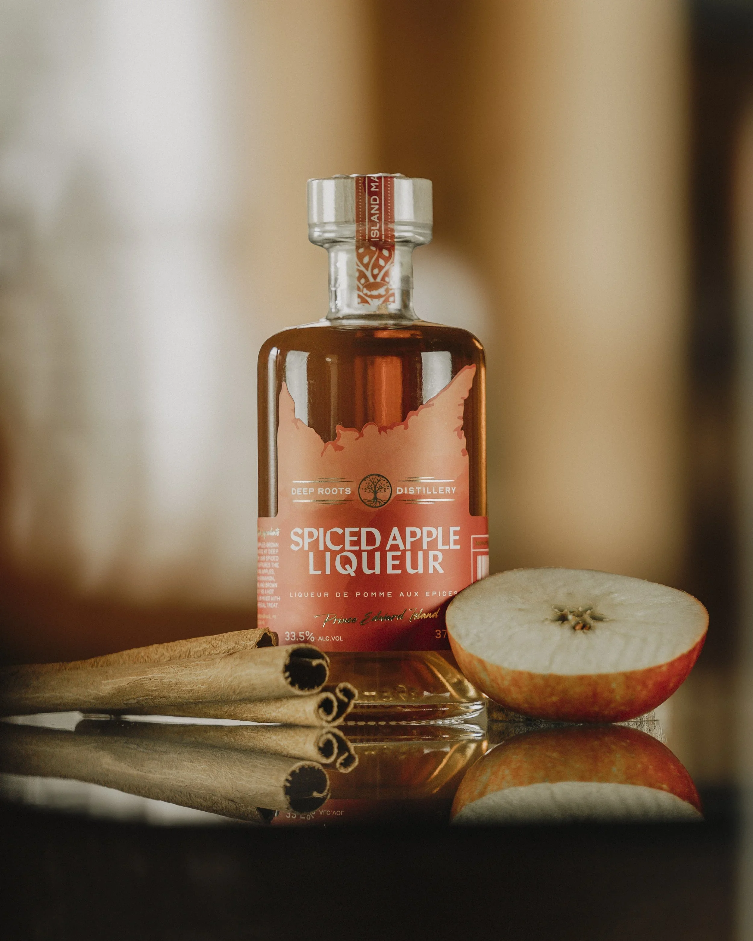 A bottle of spiced apple liqueur with a label featuring a stylized apple and a maple leaf, placed next to a halved apple and cinnamon sticks on a textured surface.