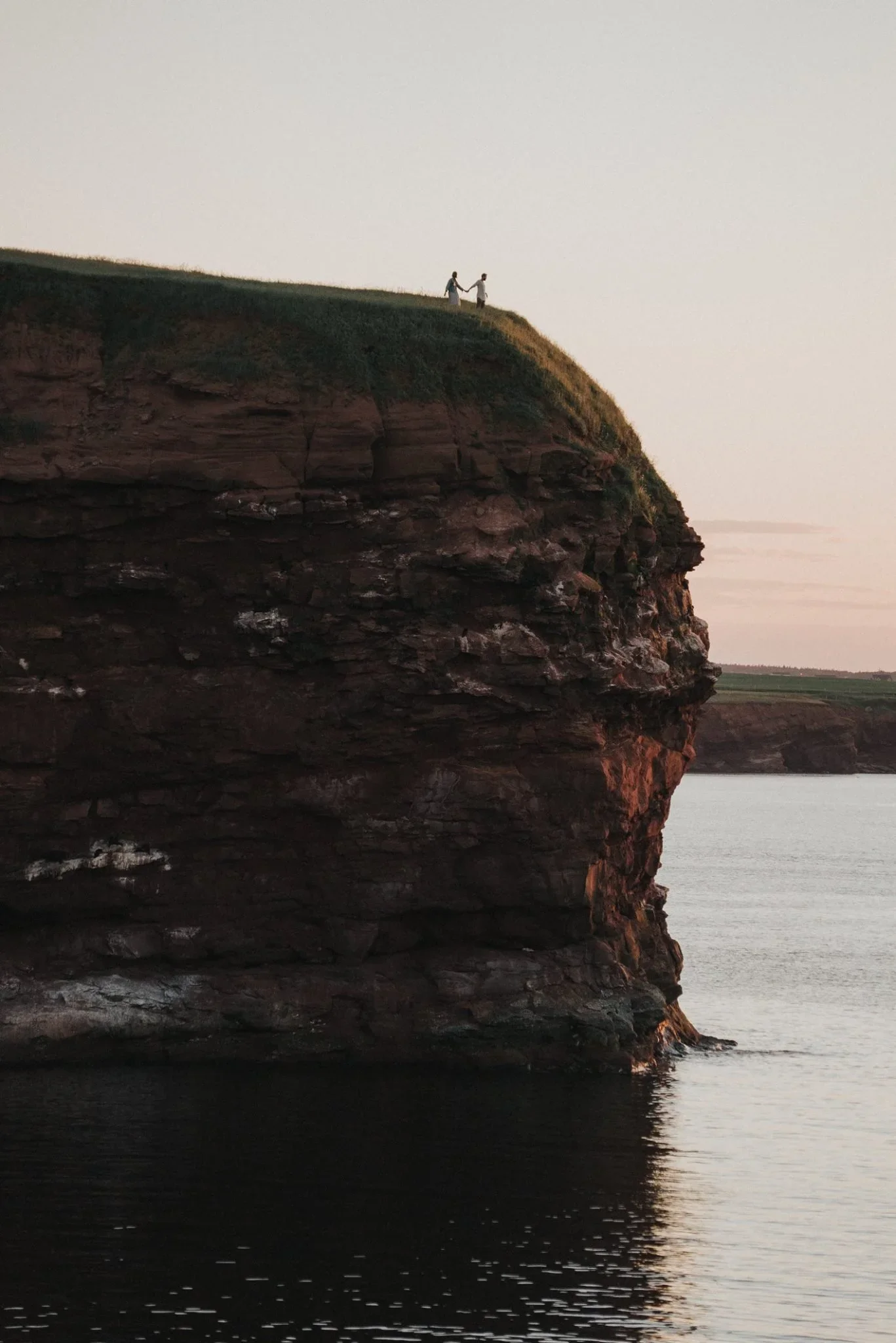 A couple holding hands walking along the grassy top of a large rocky cliff overlooking a body of water during sunset.