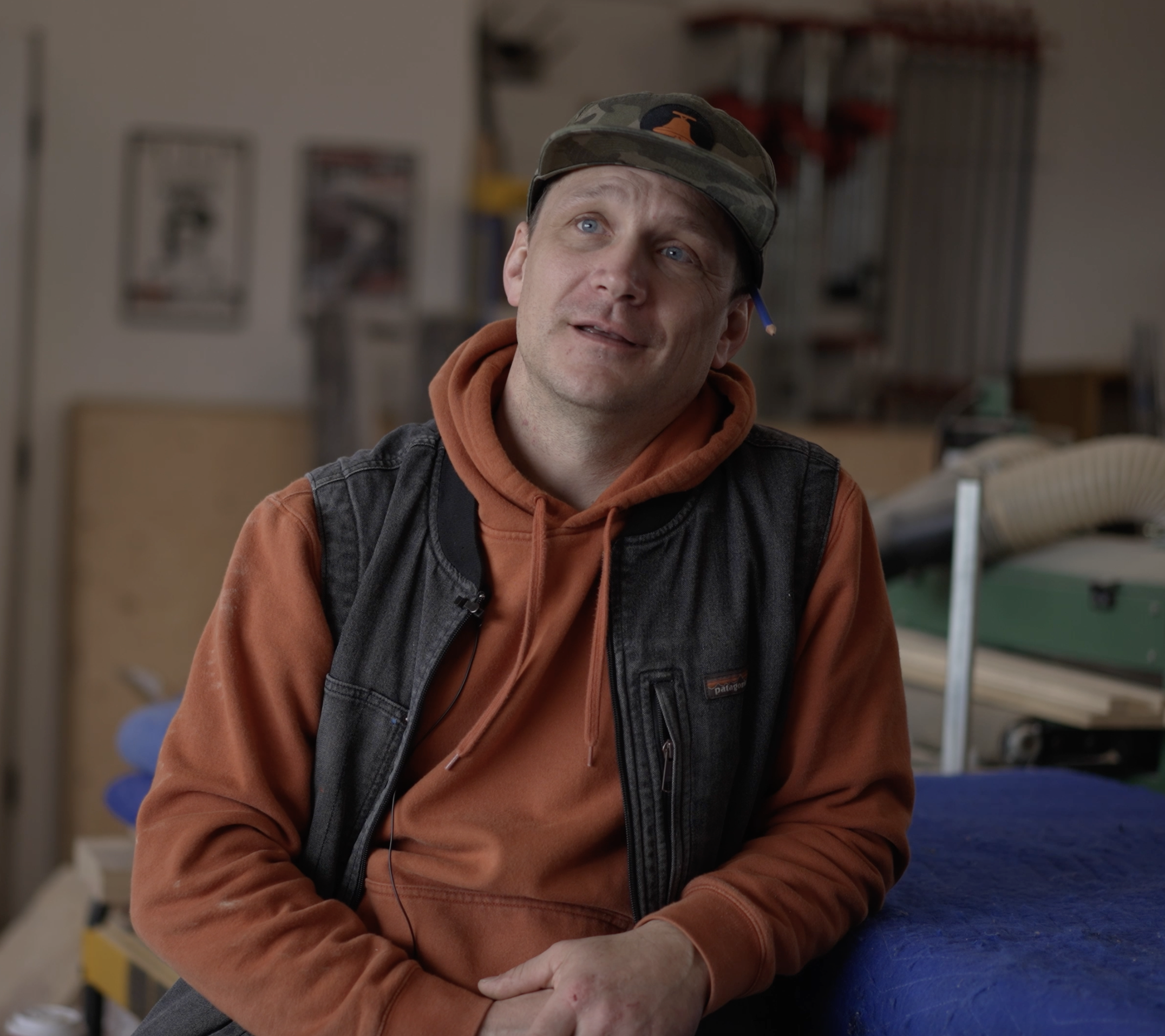 A man with light skin and blue eyes wearing a camouflage cap with an orange logo, an orange hoodie, and a black vest, sitting indoors with a neutral expression.