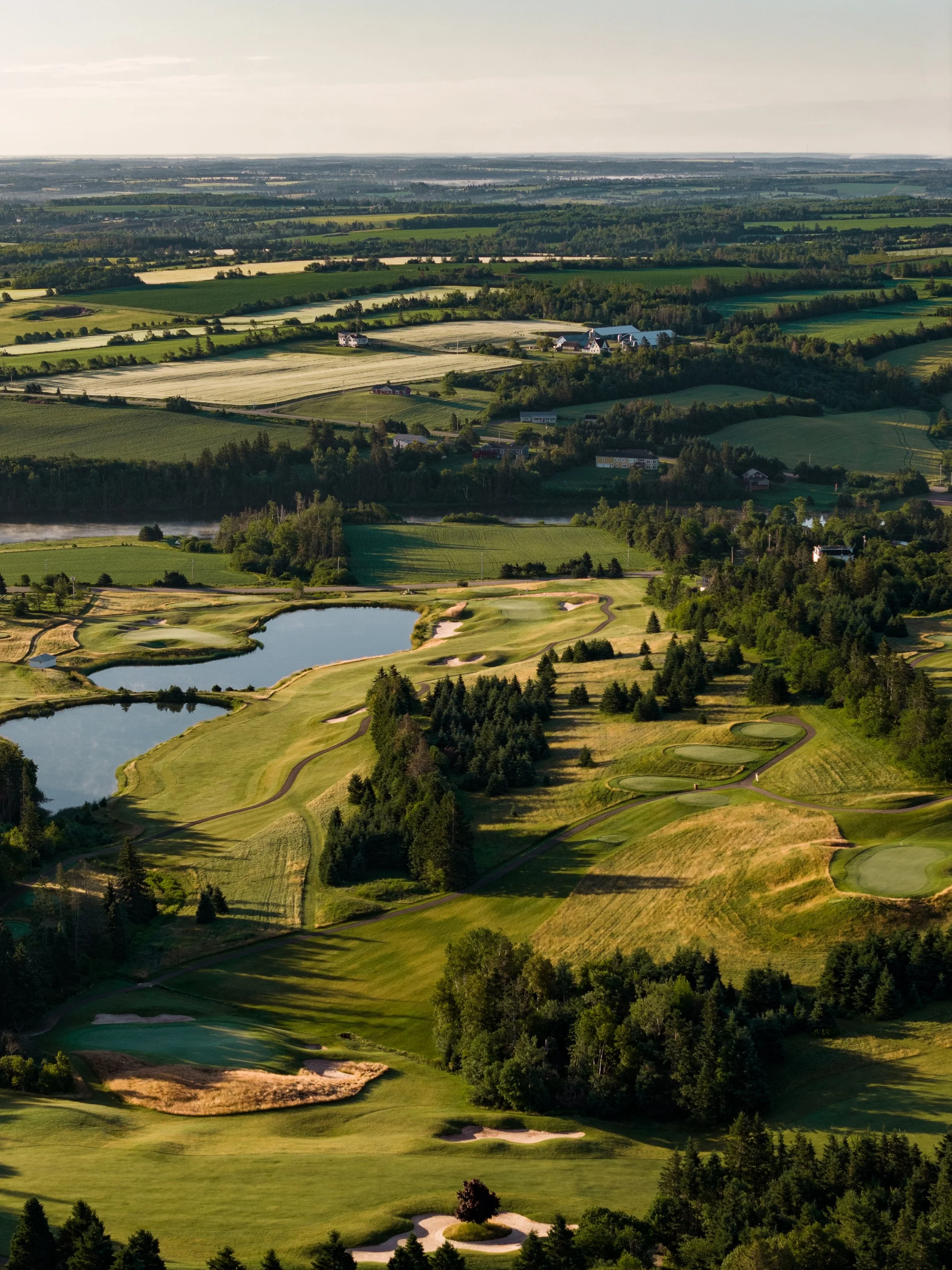 Aerial view of a golf course with water hazards, sand traps, and green fairways, surrounded by trees and residential buildings in the distance.