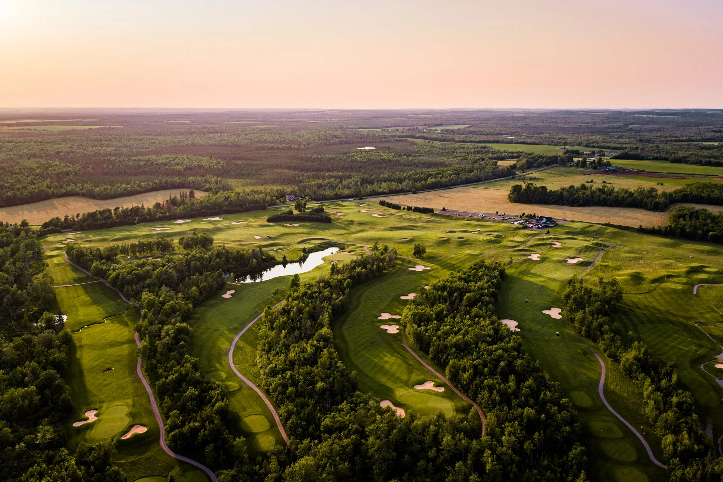 Aerial view of a golf course surrounded by trees with a parking lot and clubhouse nearby, extending into a rural landscape with farmland and forests under a pinkish sky.