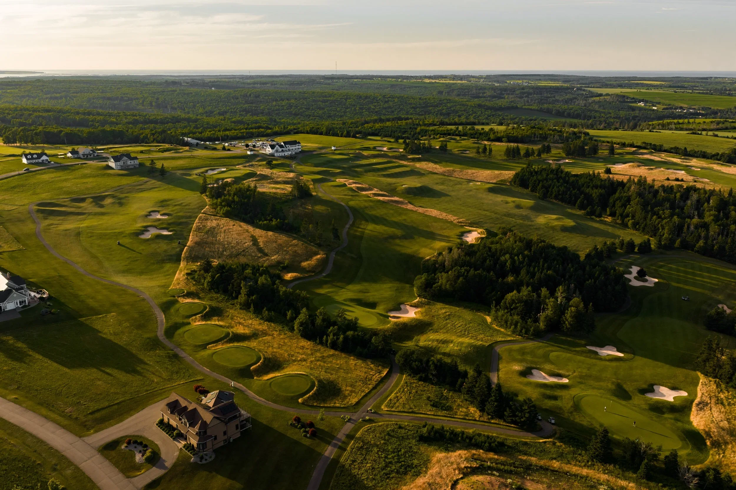 Aerial view of a golf course with lush green fairways, sand traps, and surrounding trees, with residential houses nearby and open fields in the distance.