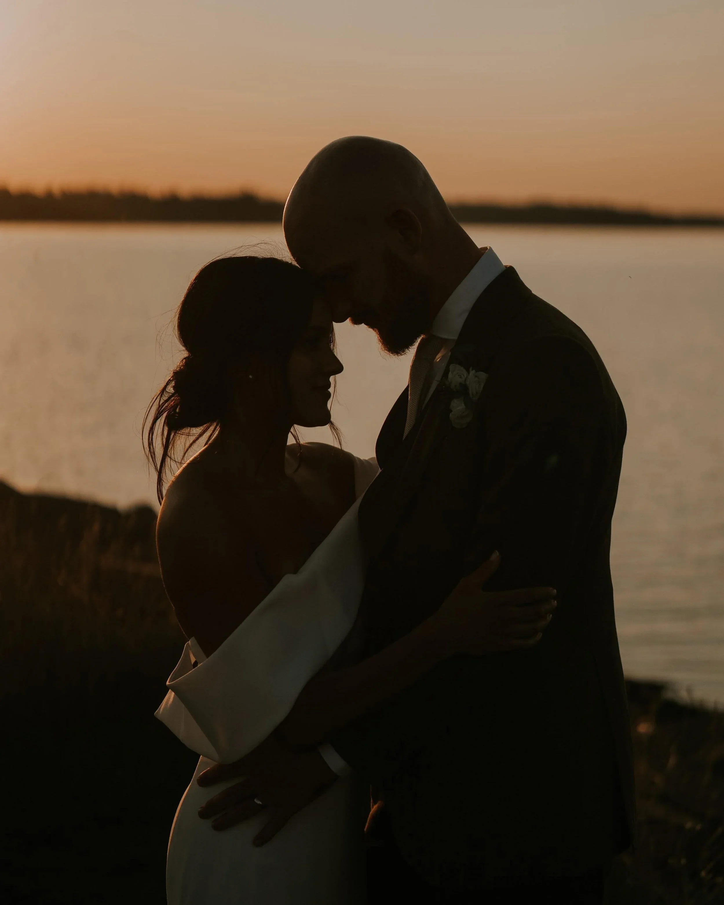 Silhouettes of a couple embracing at sunset near a body of water, with the man wearing a tuxedo and the woman in a white dress.