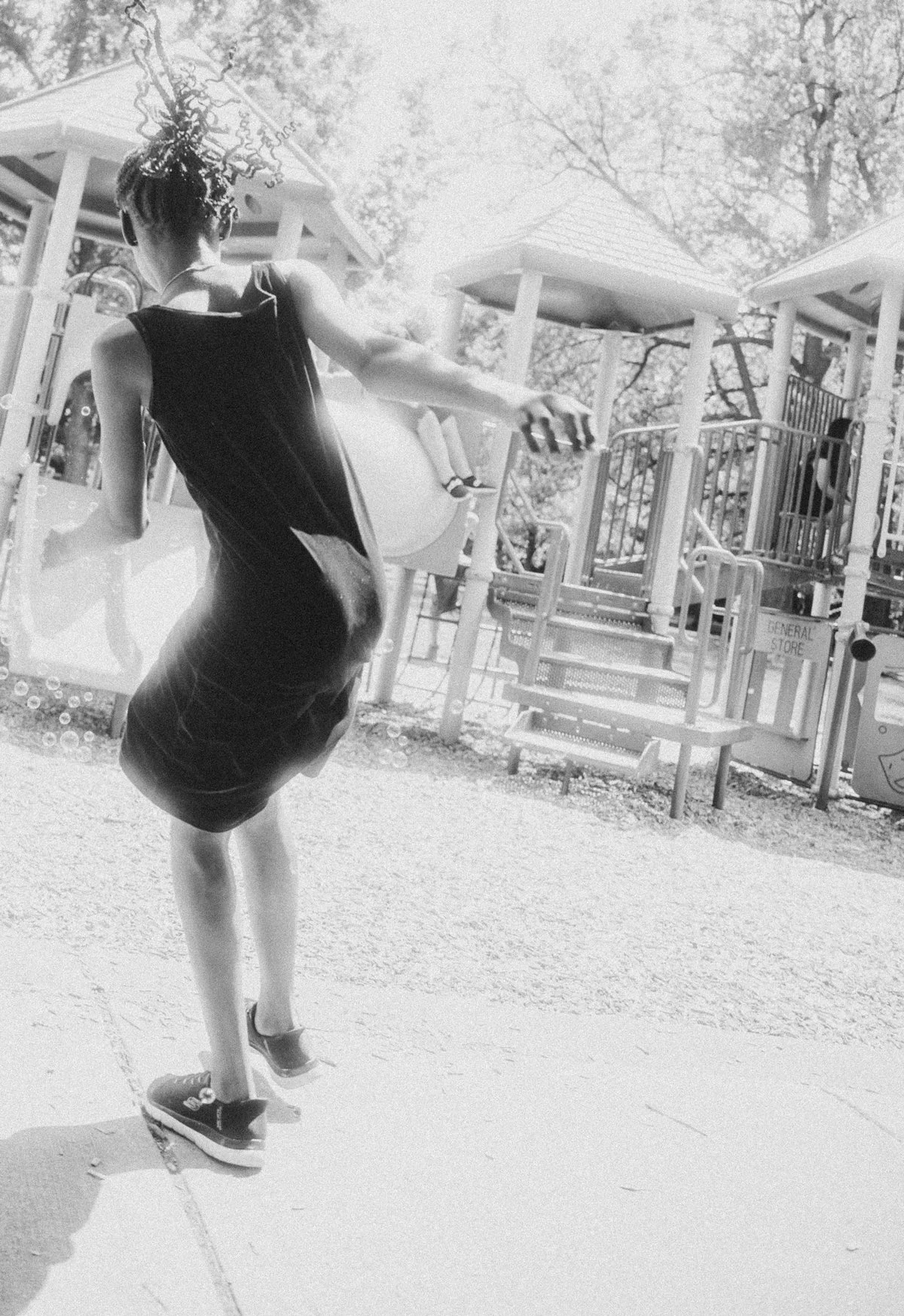 A woman in a black dress and sneakers standing outdoors near a playground, with her head turned away and her hand outstretched.