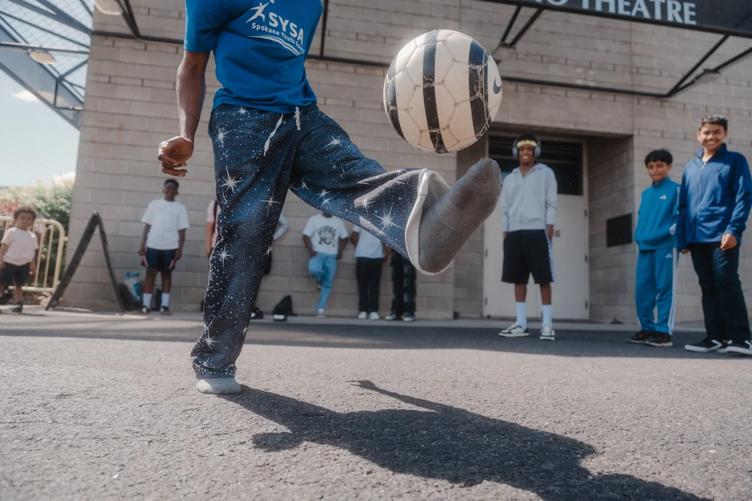 A person wearing galaxy-patterned pants and a blue shirt is performing a kick to a soccer ball. Several children are standing in the background near a building, watching the activity, with one of them smiling.
