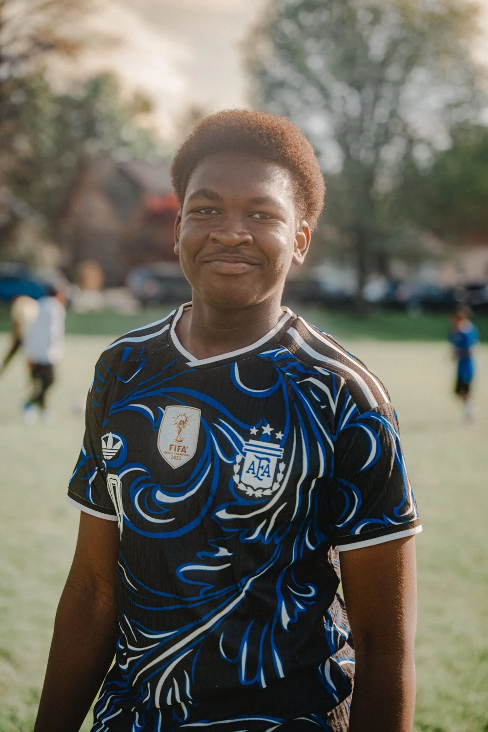Young man smiling outdoors on a soccer field, wearing an Argentina national team jersey with FIFA and Adidas badges, with blurred background of other players and trees.
