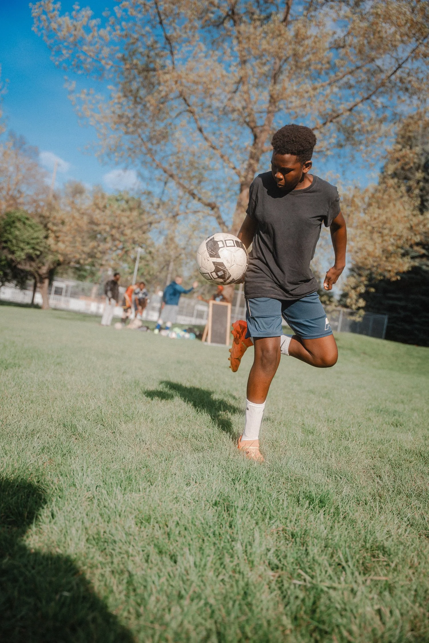 A young man wearing a black T-shirt and blue shorts playing soccer outdoors on a grassy field during daytime, with other people and trees in the background and a clear blue sky.