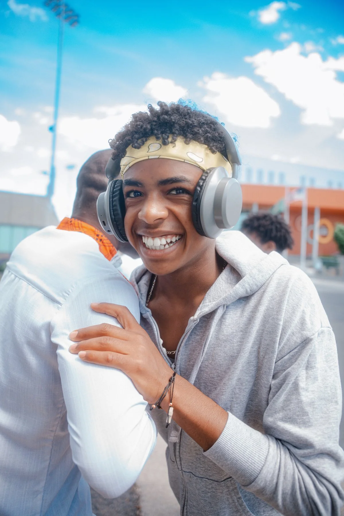 Young woman smiling and embracing someone outdoors, wearing headphones and a hoodie, with a blue sky and clouds in the background.