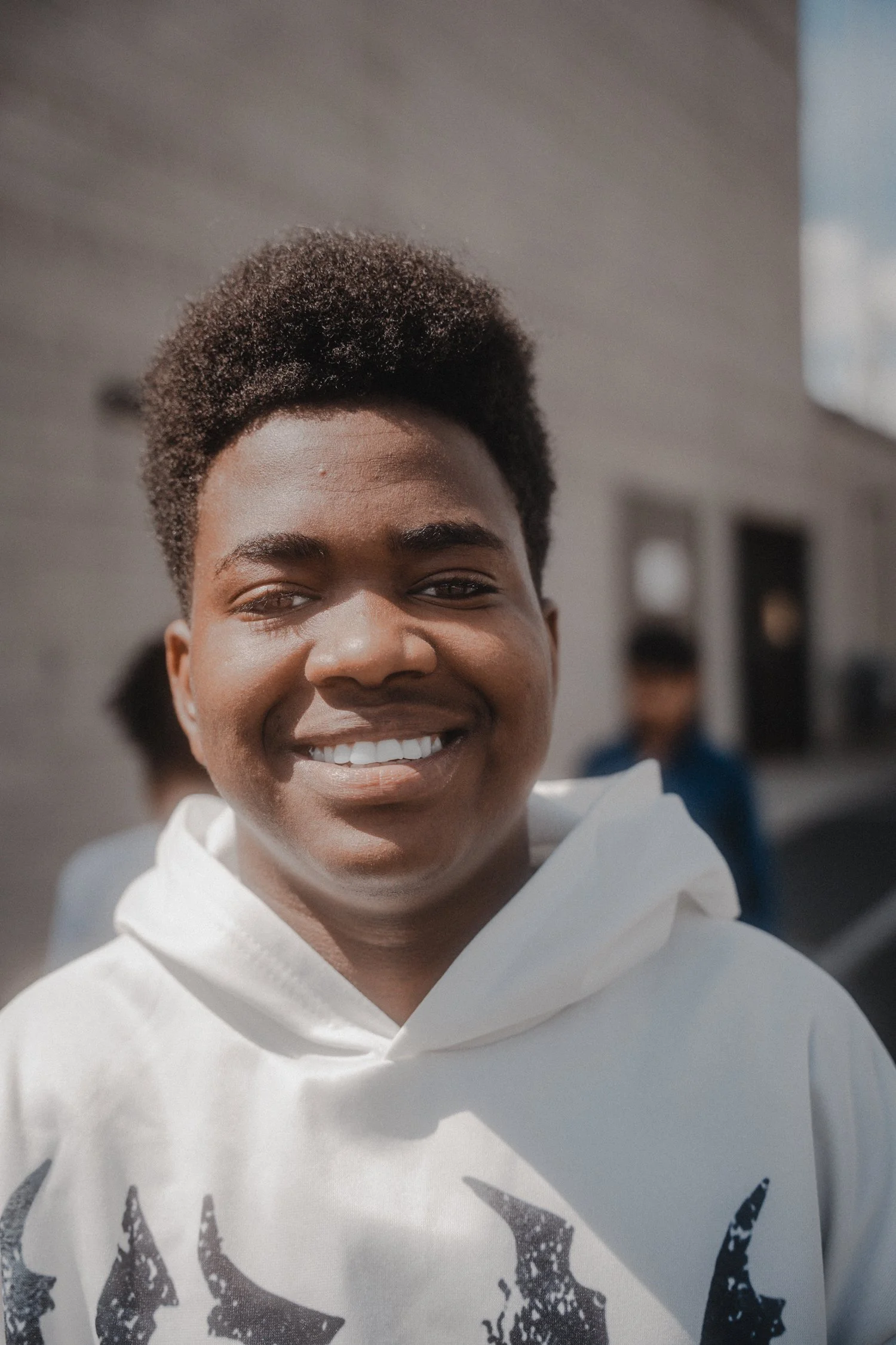 Close-up of a smiling person with short, curly hair wearing a white hoodie with black graphic print, with a blurred background featuring a building and a person.
