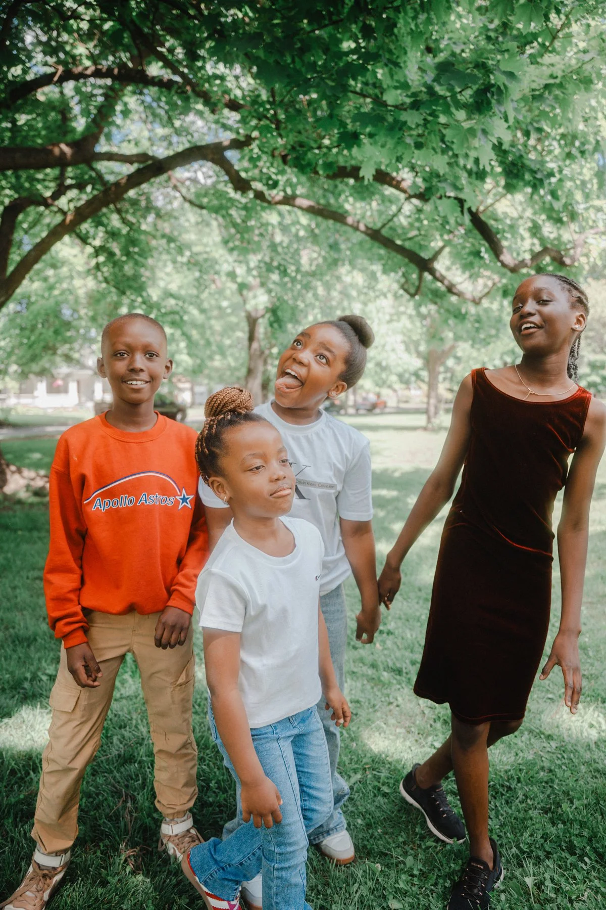 A group of four children and one woman standing outdoors under a large tree on a grassy area, enjoying a sunny day. The children are smiling and making playful expressions.