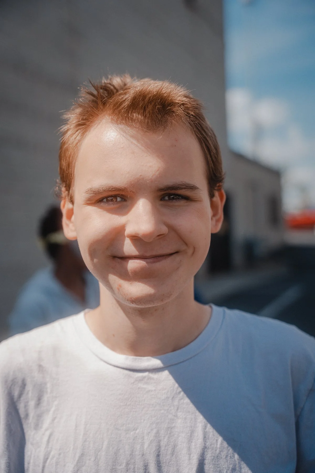 Close-up of a young man with short red hair, smiling outdoors on a sunny day, with a blue sky and people in the background.