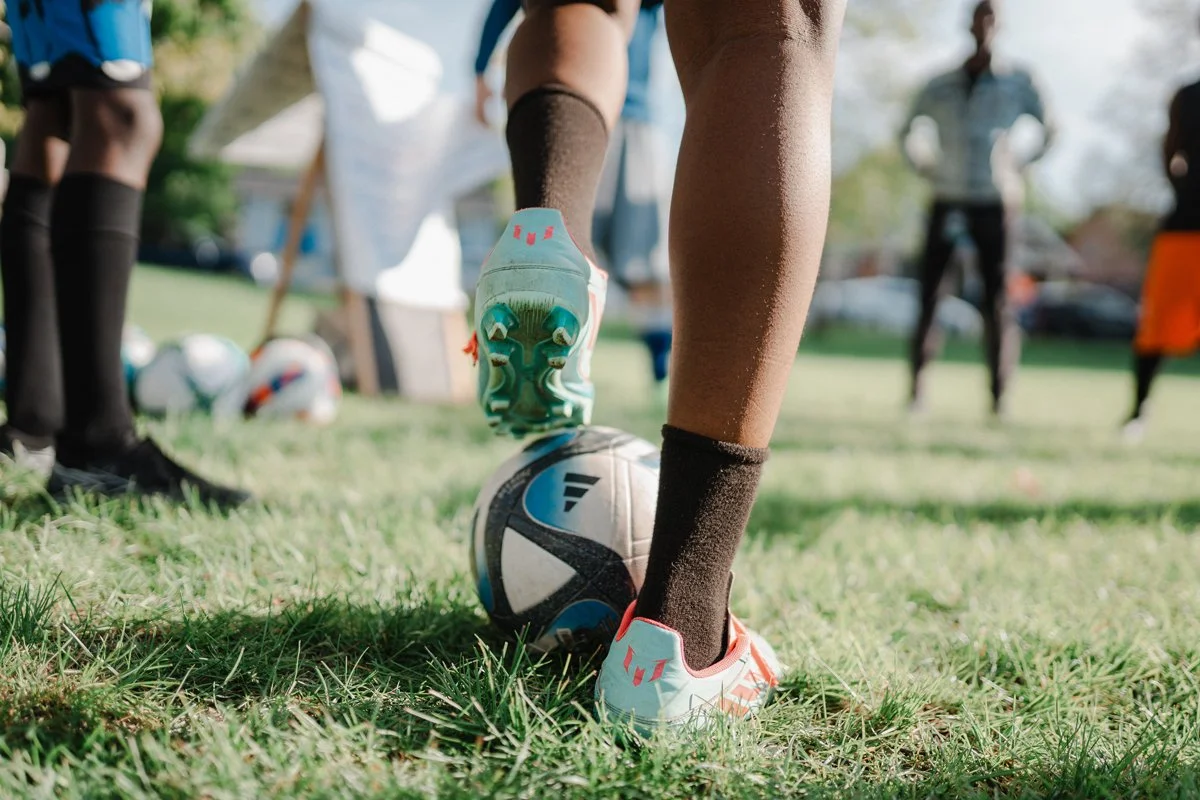Person preparing to kick a soccer ball on a grassy field during a soccer practice, with other players and coaches in the background.