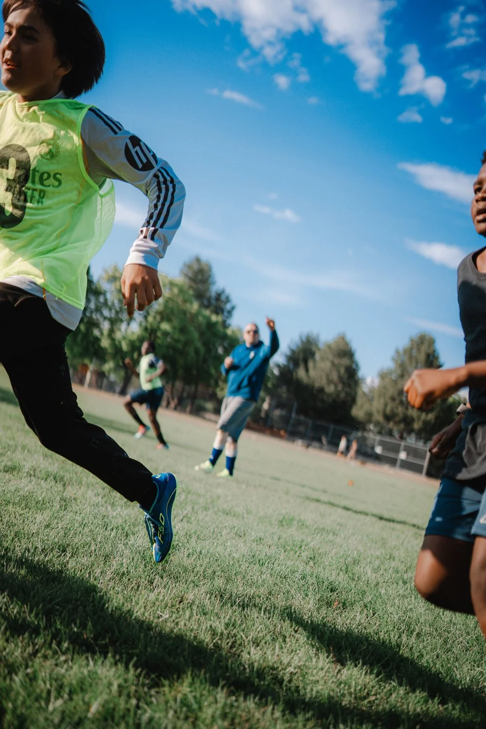 Children playing soccer on a grassy field under a blue sky with some clouds, with a coach or adult cheering in the background.