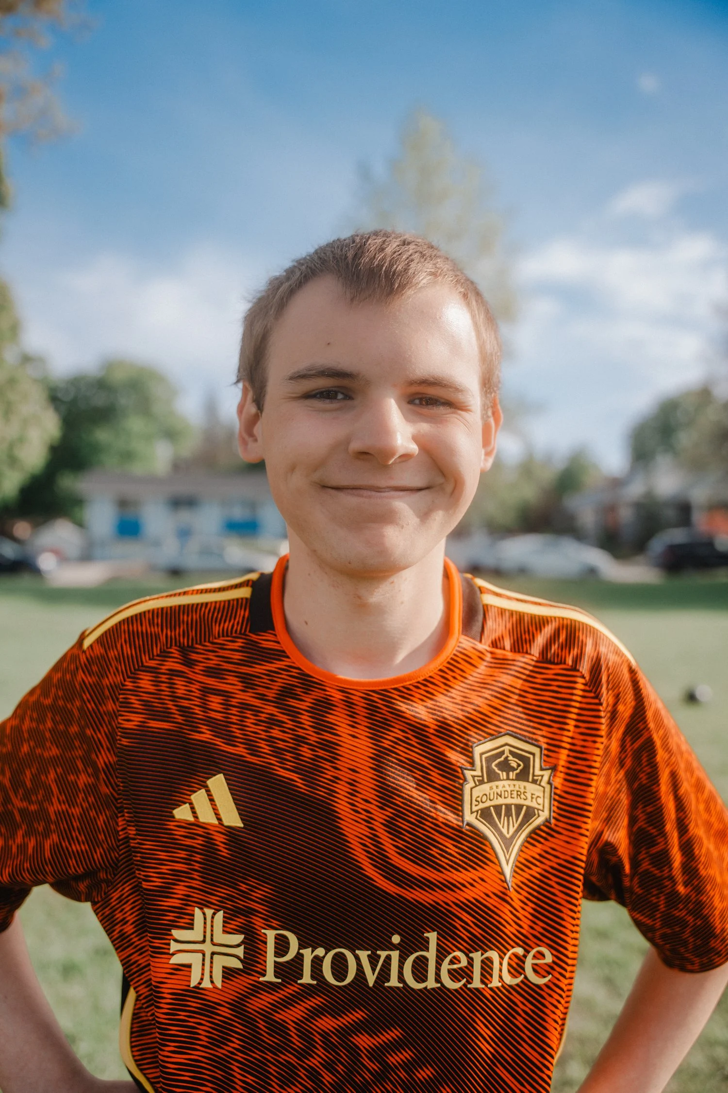 A young man with short light brown hair and fair skin smiling outdoors on a sunny day, wearing an orange and black sports jersey with a Providence Souders FC logo and Adidas branding, standing in front of a background with trees and houses.