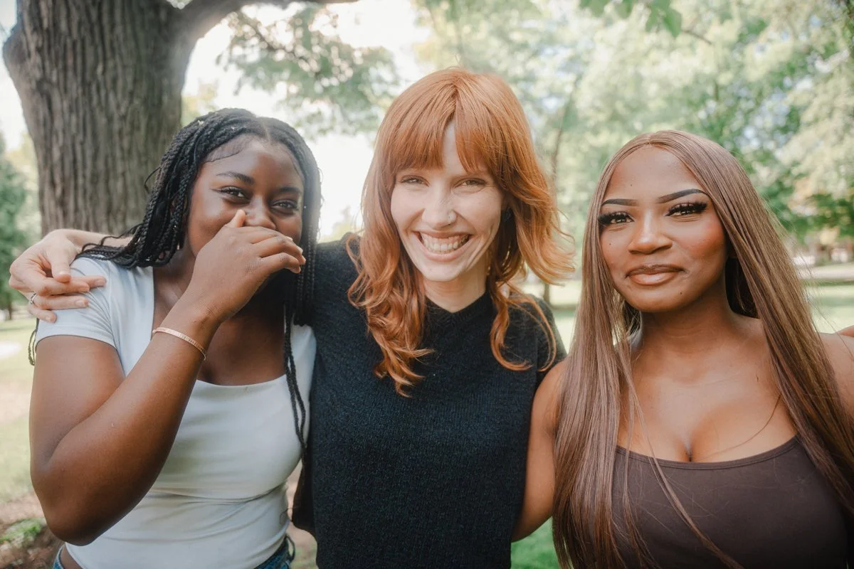 Three women smiling and hugging outdoors in a park with trees in the background.