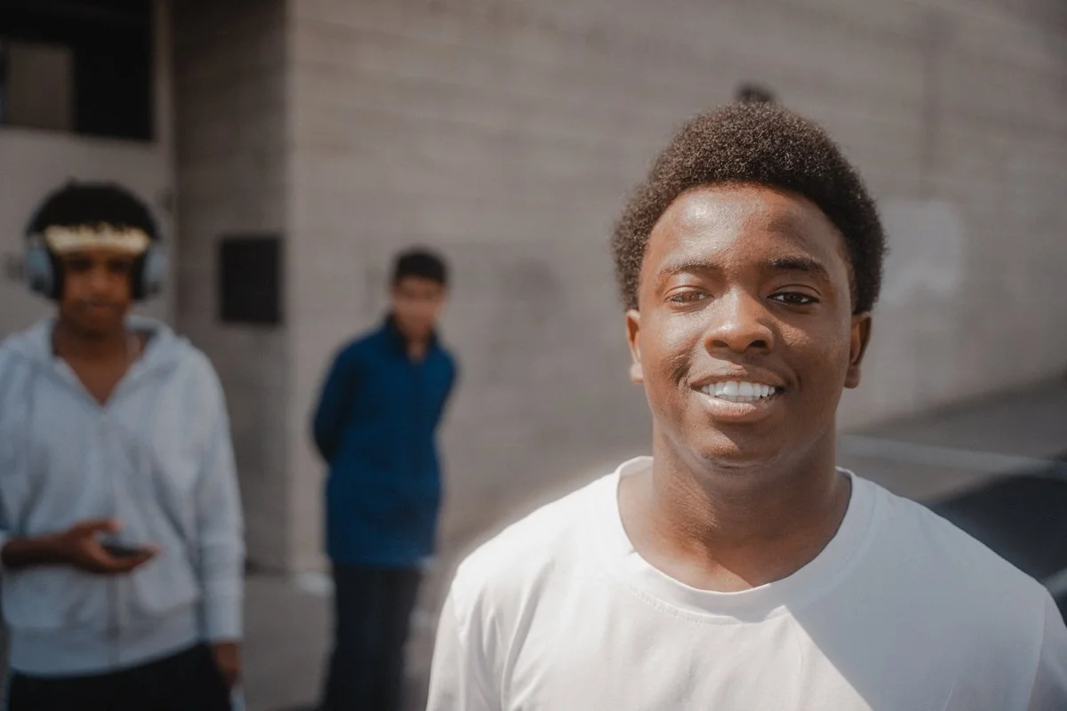 Young man smiling in front of a gray building, with two people in the background, one wearing headphones and holding a phone, and the other standing with arms crossed.