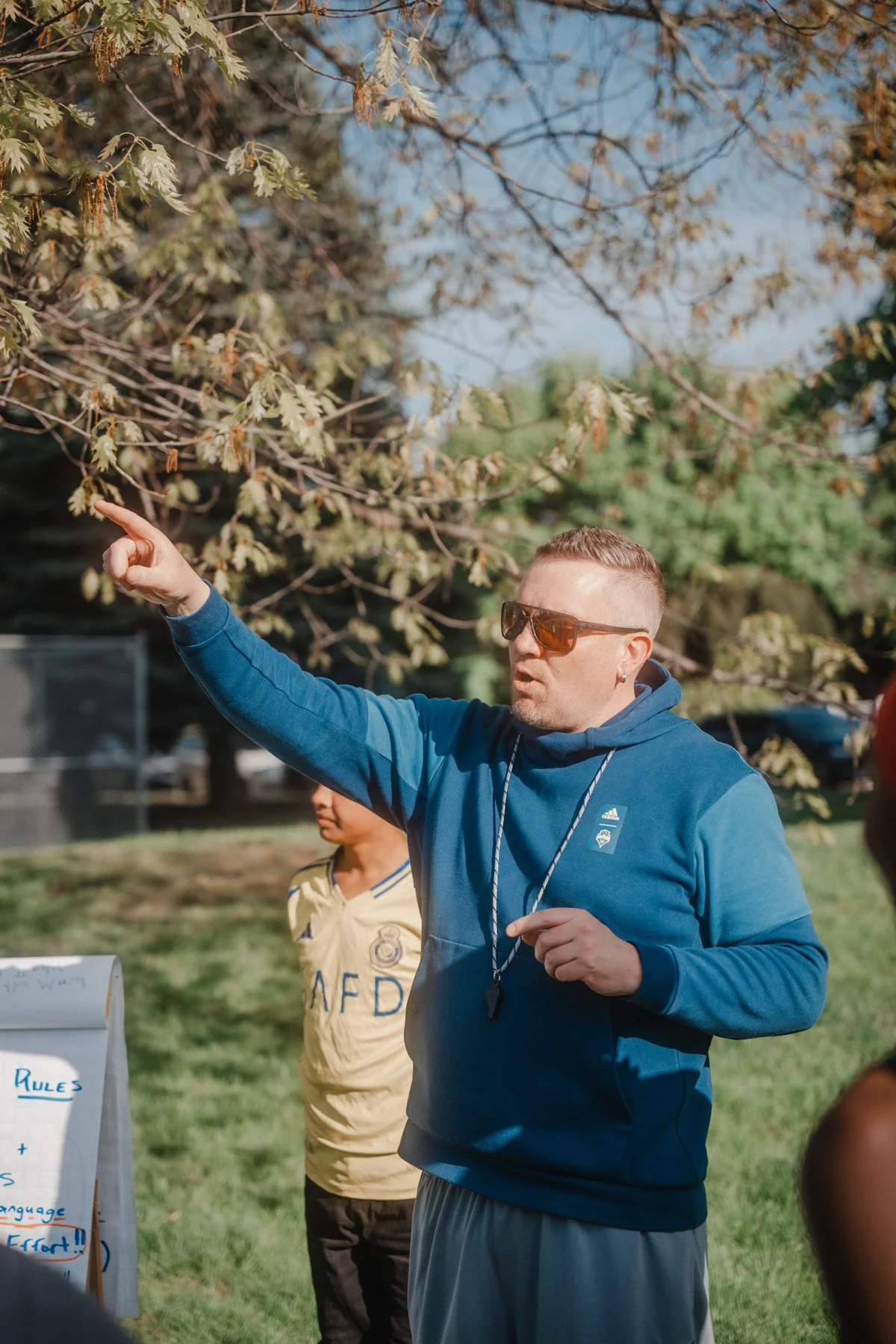 A man wearing sunglasses and a blue hoodie is pointing at a tree during an outdoor event, with a young boy in a yellow jersey standing nearby.