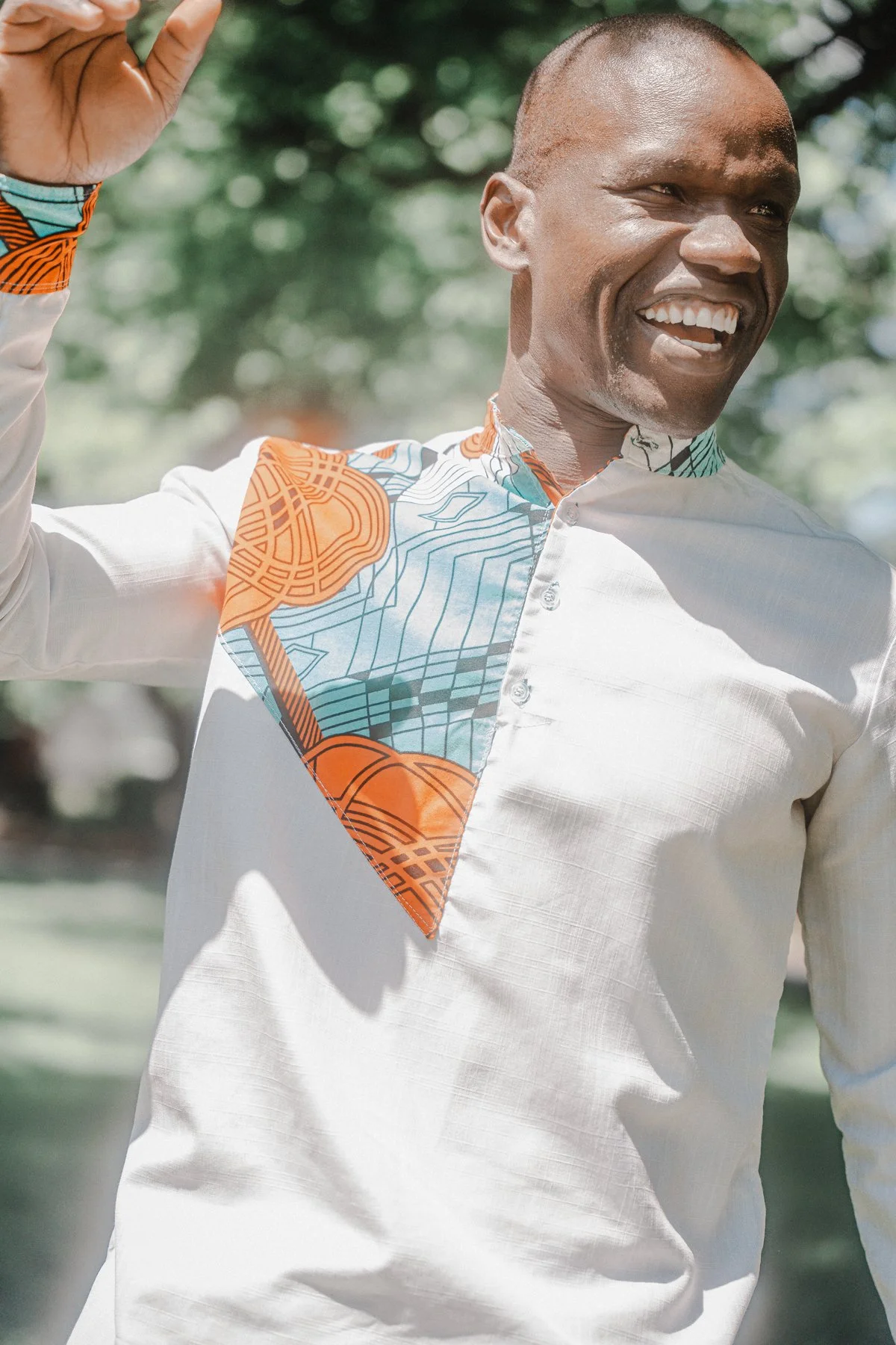 A smiling man dressed in a white shirt with a colorful patterned fabric draped over his shoulder, outdoors with blurred green trees in the background.