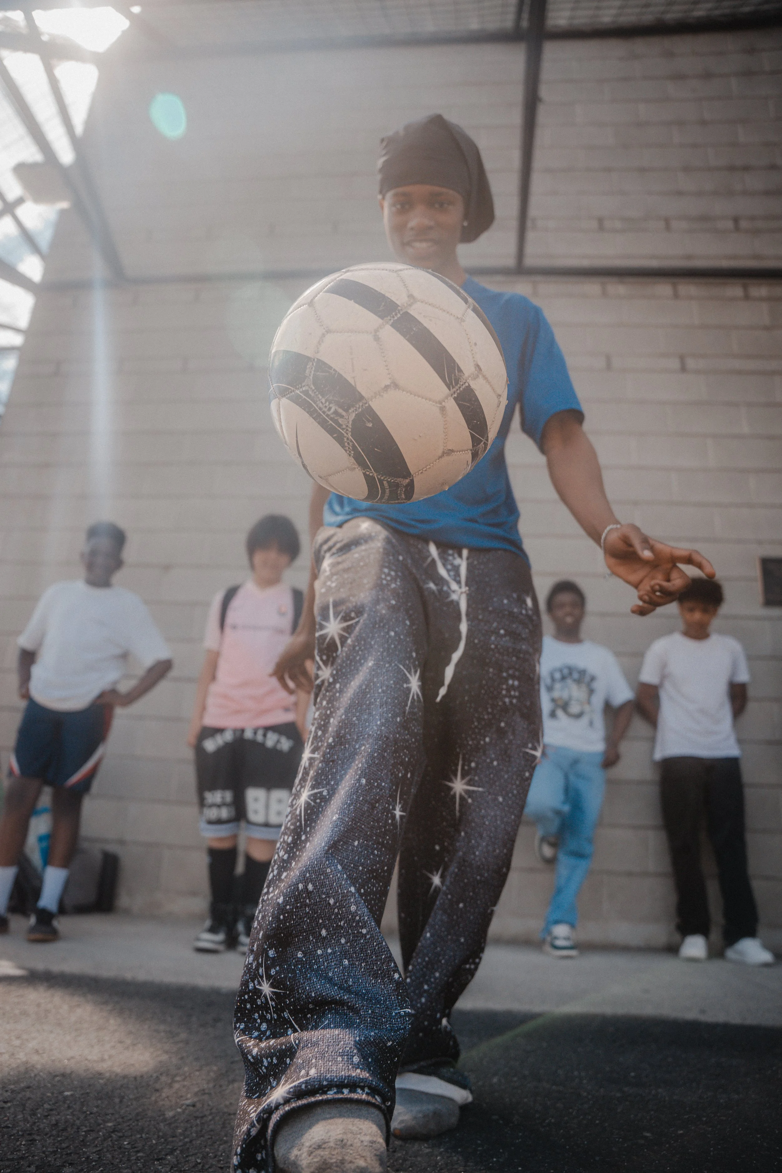 A person wearing galaxy-print pants is performing a soccer trick with a soccer ball in an outdoor area. Several children are watching in the background, standing against a brick wall. The person is wearing a black headwrap, a blue shirt, and white sh