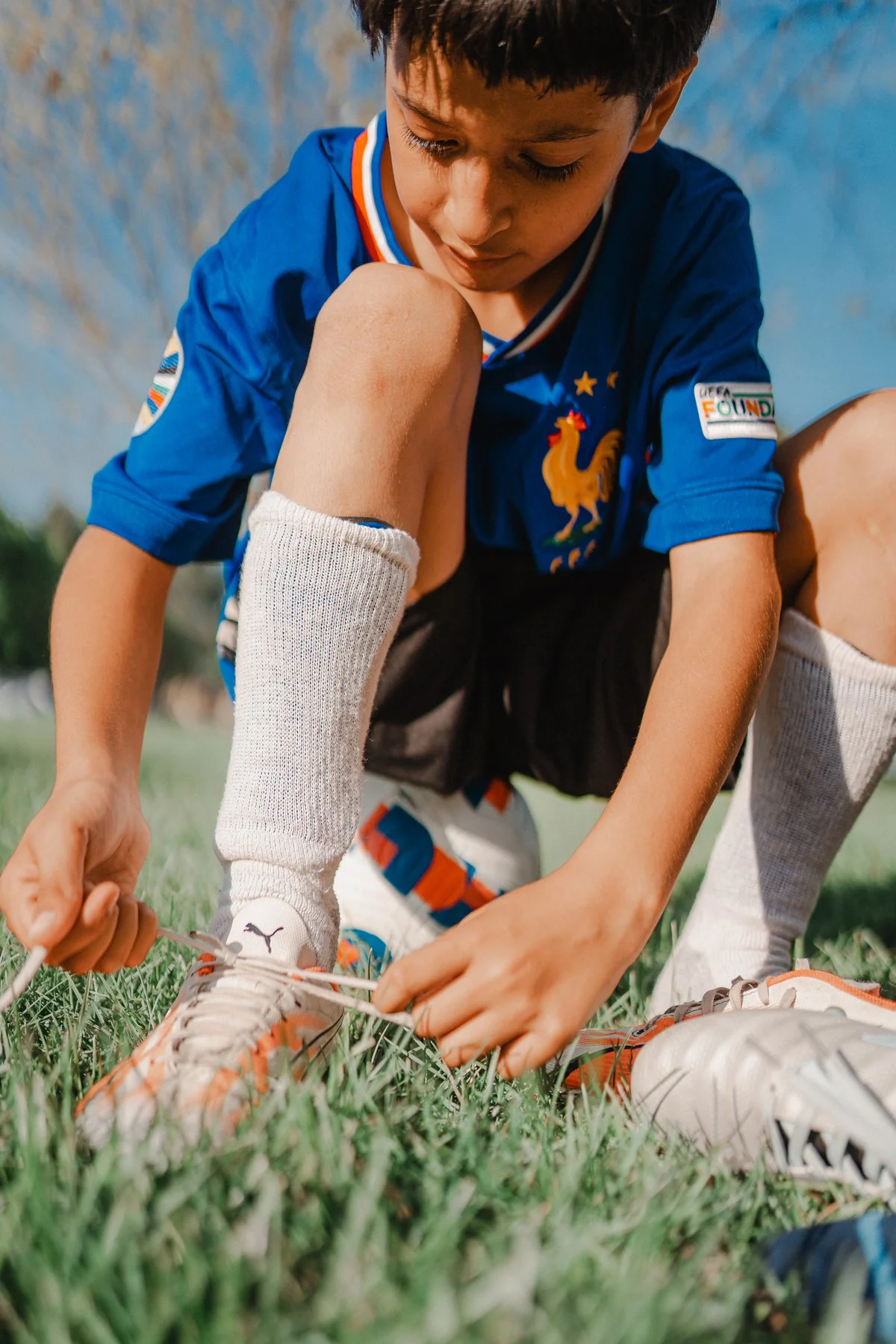 A young boy tying his shoelaces on a grassy field, wearing a blue soccer jersey with patches and a lion emblem, white socks, and athletic shoes. The background includes a blue sky and some blurred trees.