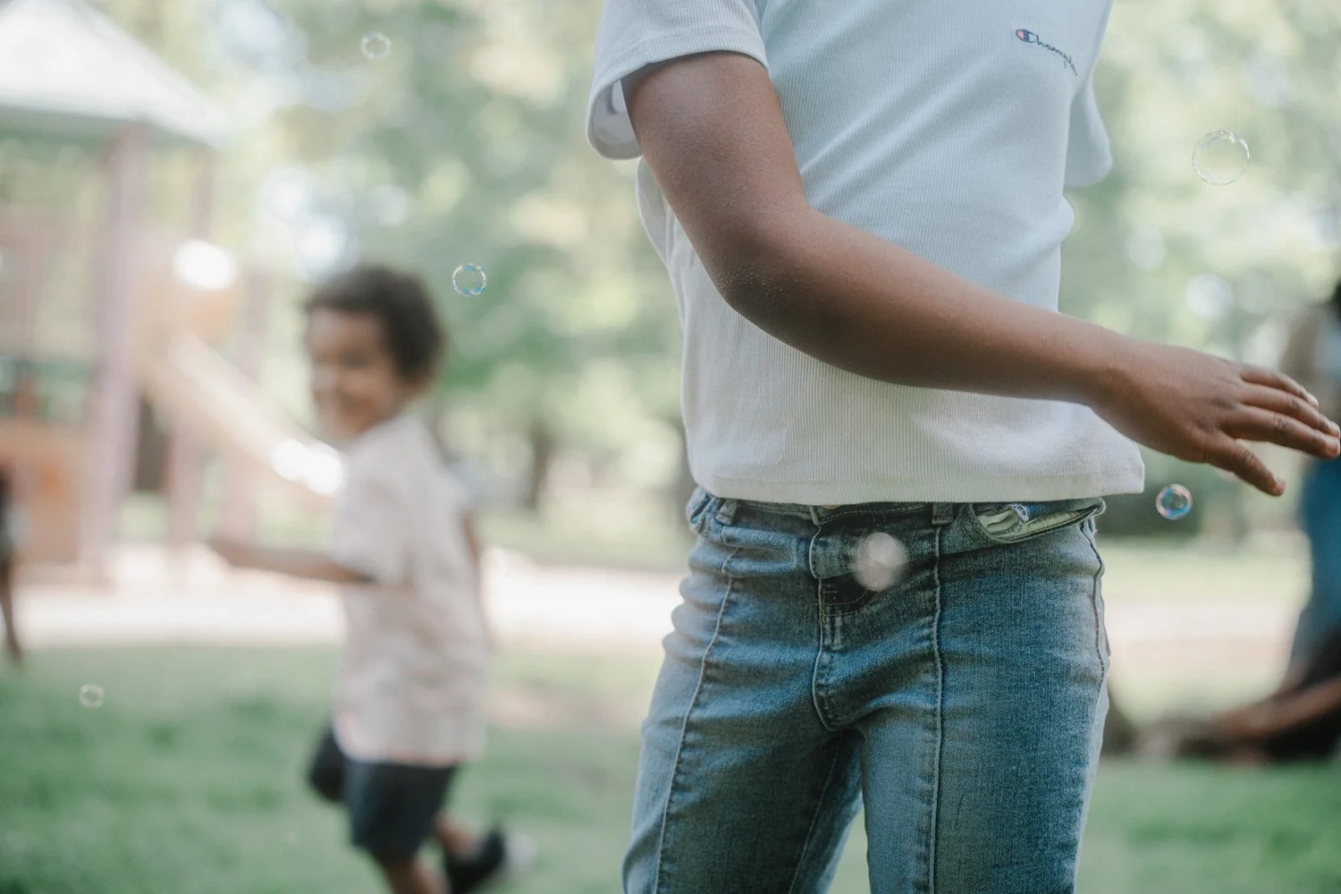 Teen girl in a grey T-shirt and jeans playing with bubbles outdoors in a park, with blurred background of trees and another girl in the background.