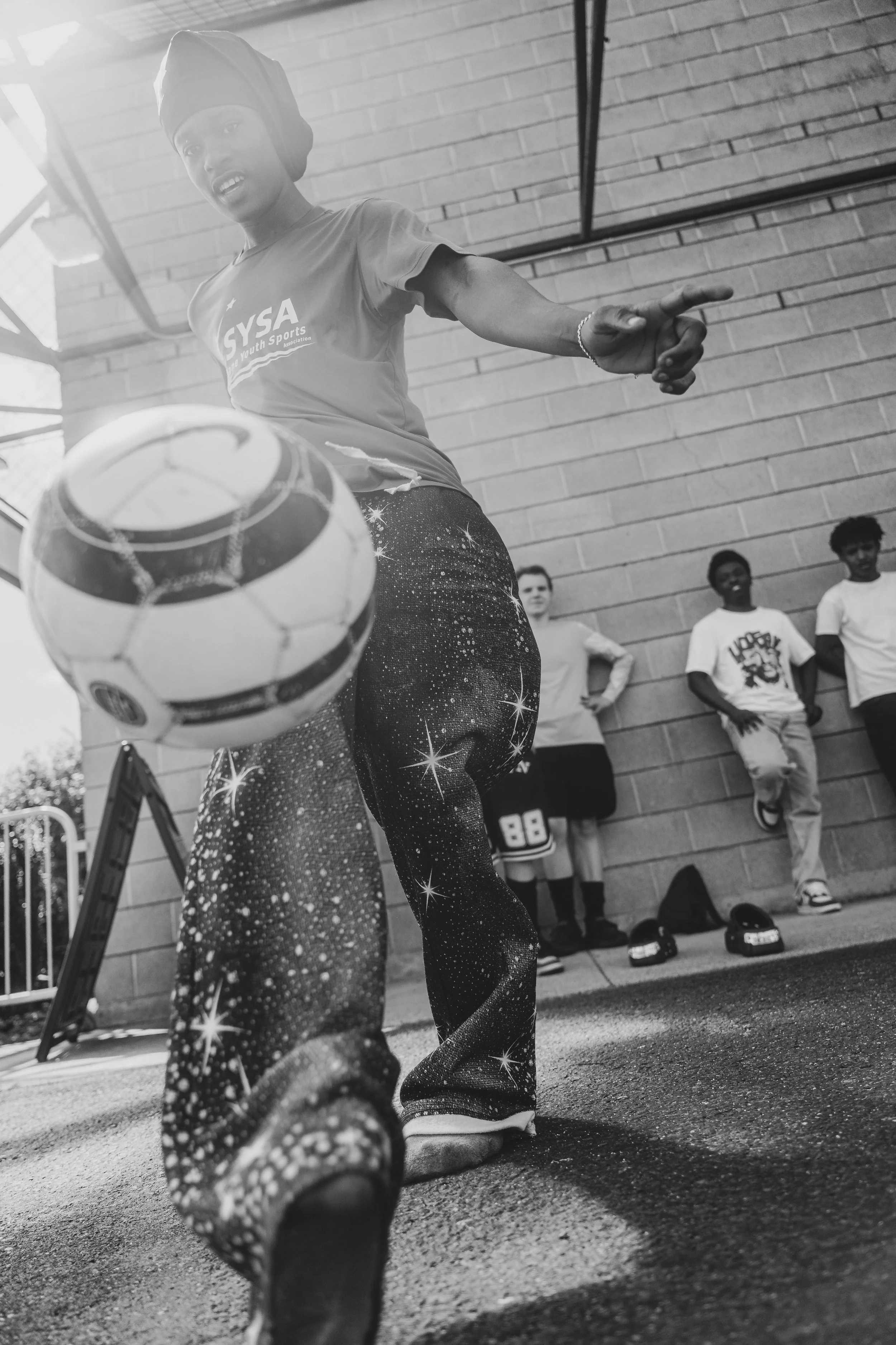 A woman wearing galaxy-print pants and a shirt with the logo 'GYSA Youth Sports' is kicking a soccer ball indoors. She is wearing a headwrap and bracelets. Several people stand against a brick wall in the background.