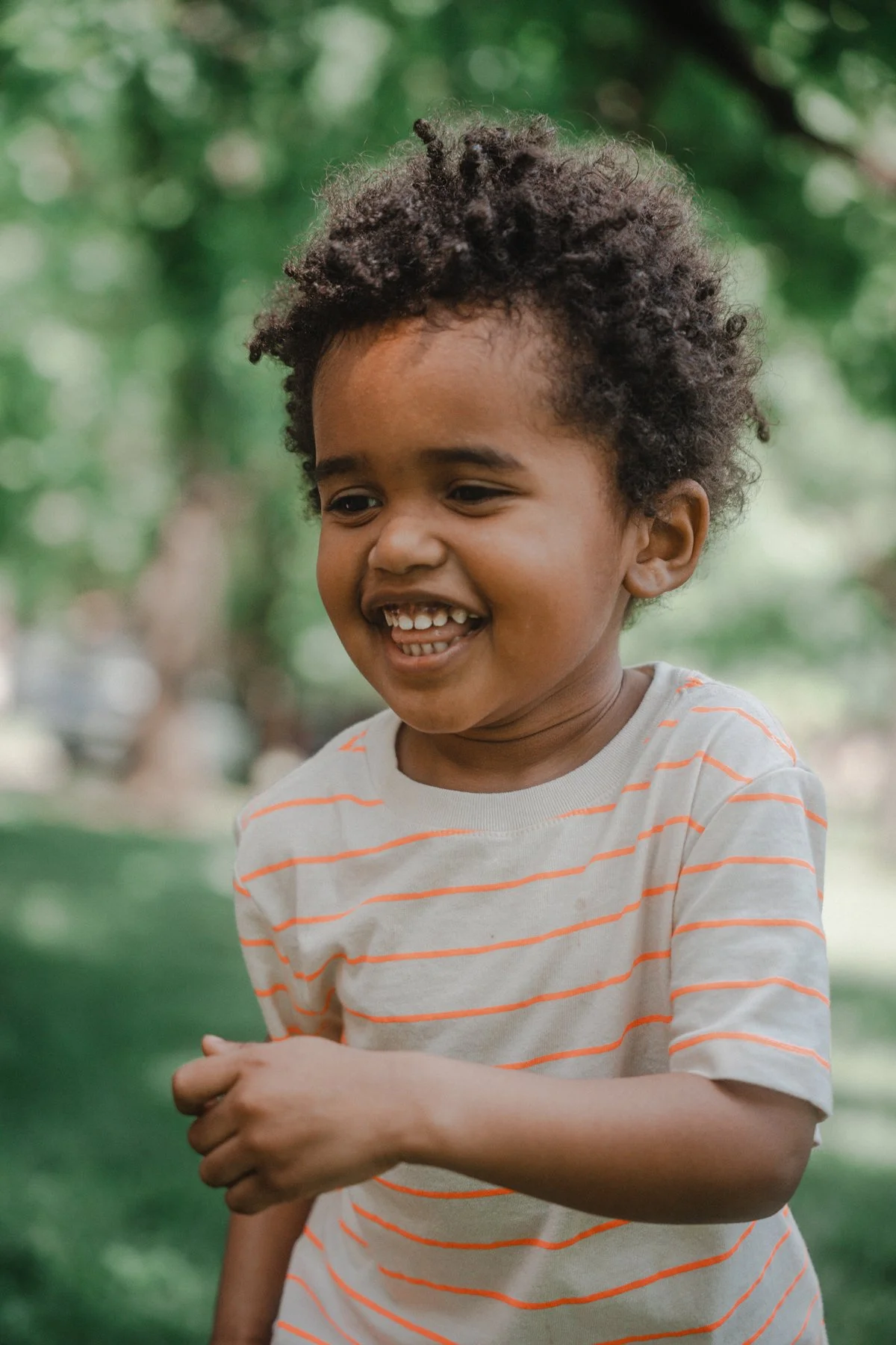 A young smiling boy with curly hair wearing a striped t-shirt outdoors with green trees in the background.