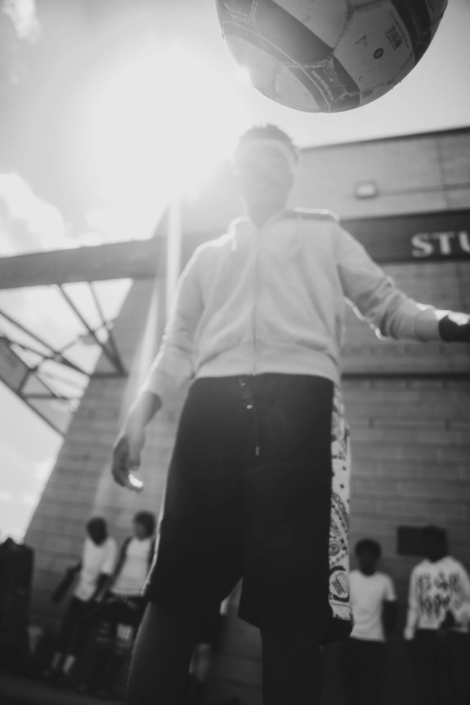 A person standing outdoors, with the sun shining brightly behind them. The person is wearing a hoodie and dark pants, and their face is not clearly visible. In the background, there are several other people, some sitting on steps, a building, and a c