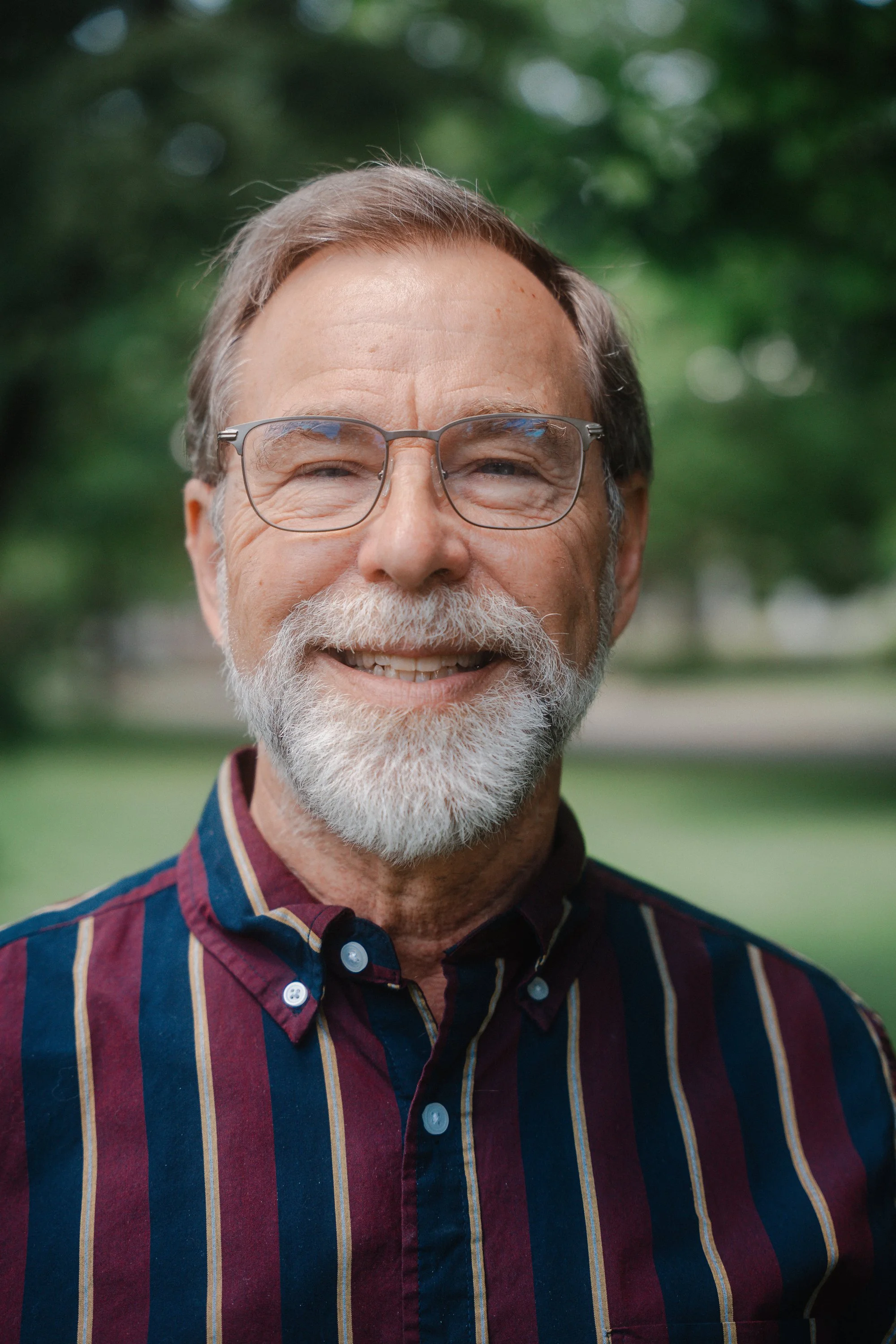 Close-up of an elderly man smiling outdoors, wearing glasses and a striped button-up shirt, with a blurred green background.