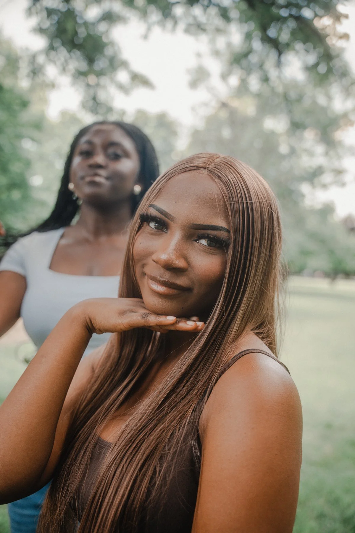 Two women outdoors, one with long, straight, brown hair, smiling and resting her chin on her hand, and the other with dark, curly hair, standing behind her, slightly out of focus, in a park-like setting with trees in the background.