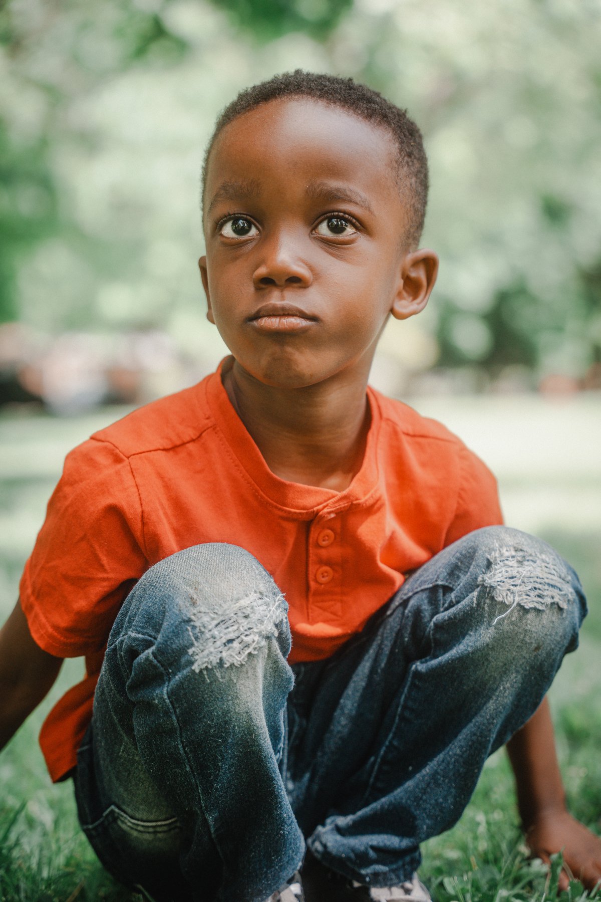 A young boy with short curly hair, wearing an orange shirt and ripped jeans, sitting outdoors on grass with a blurred green background.