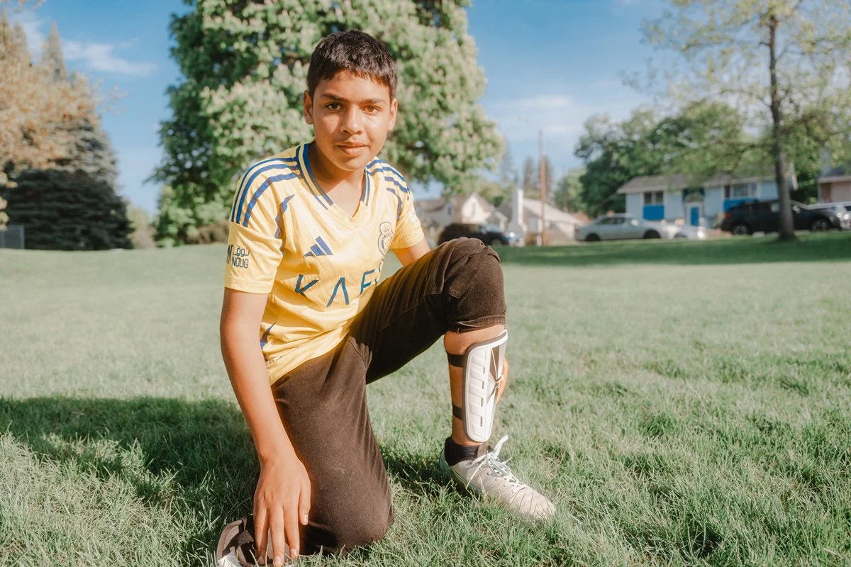 A young boy kneeling on the grass in a park with one hand on the ground and the other on his knee, wearing sports attire including a yellow jersey, black pants, and a knee brace, with trees and houses in the background.
