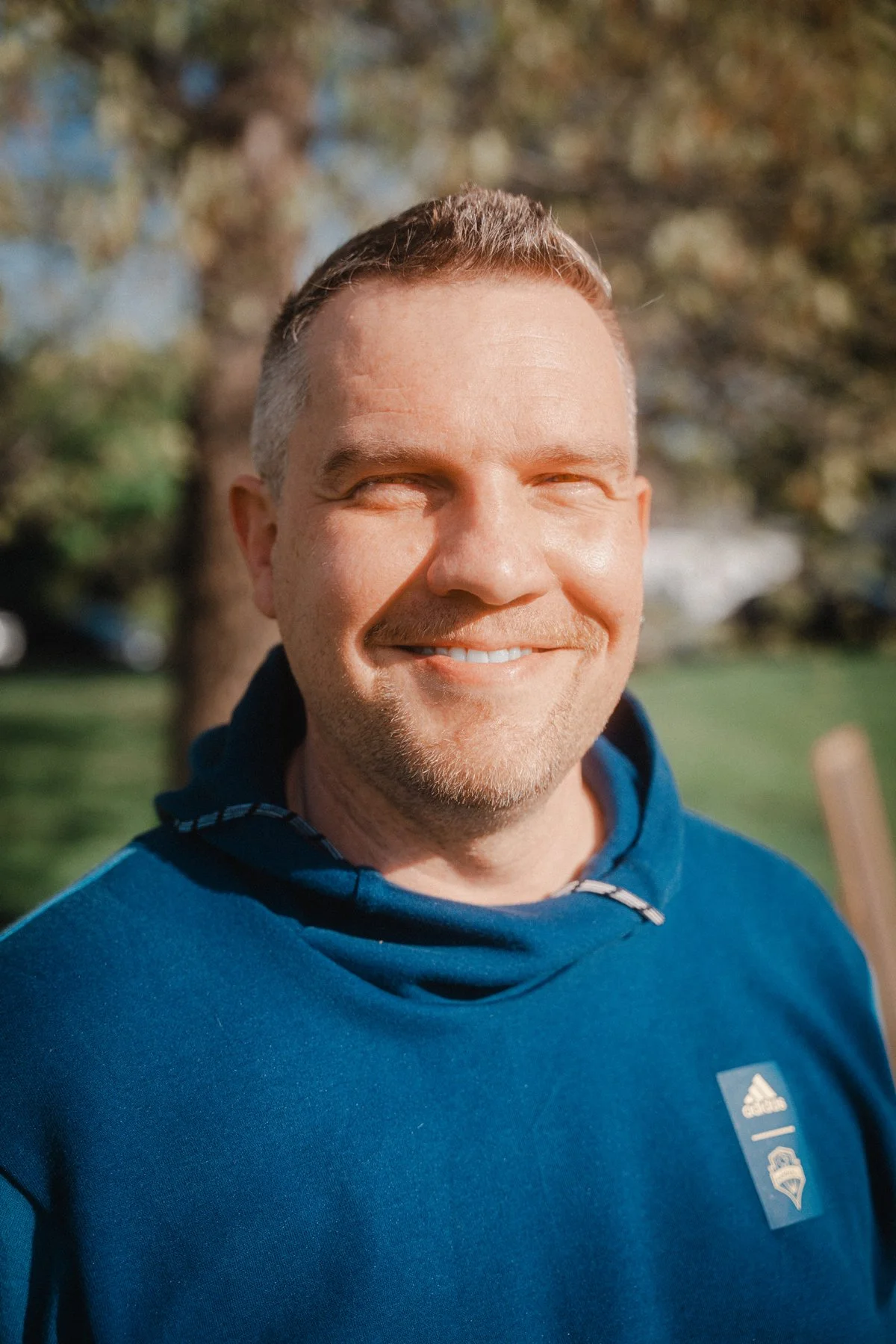 A smiling man with short reddish-brown hair, facial hair, and closed eyes, wearing a blue hoodie outdoors with trees and grass in the background.