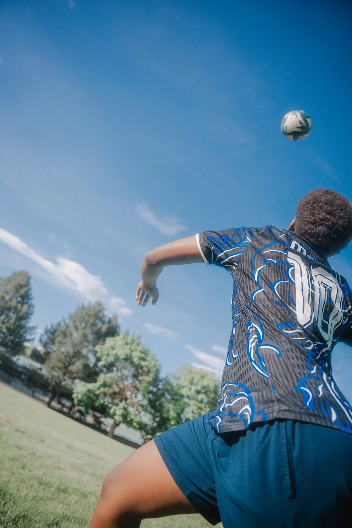 A person in a sports outfit playing soccer outdoors on a sunny day, with trees and a clear blue sky in the background.